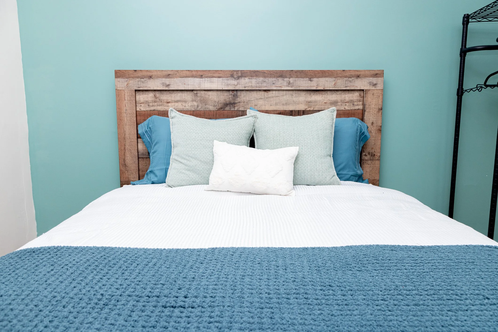 A neatly made bed with white sheets, topped with three pillows in pastel green, blue, and white with a textured pattern, against a teal-colored wall and a rustic wooden headboard.