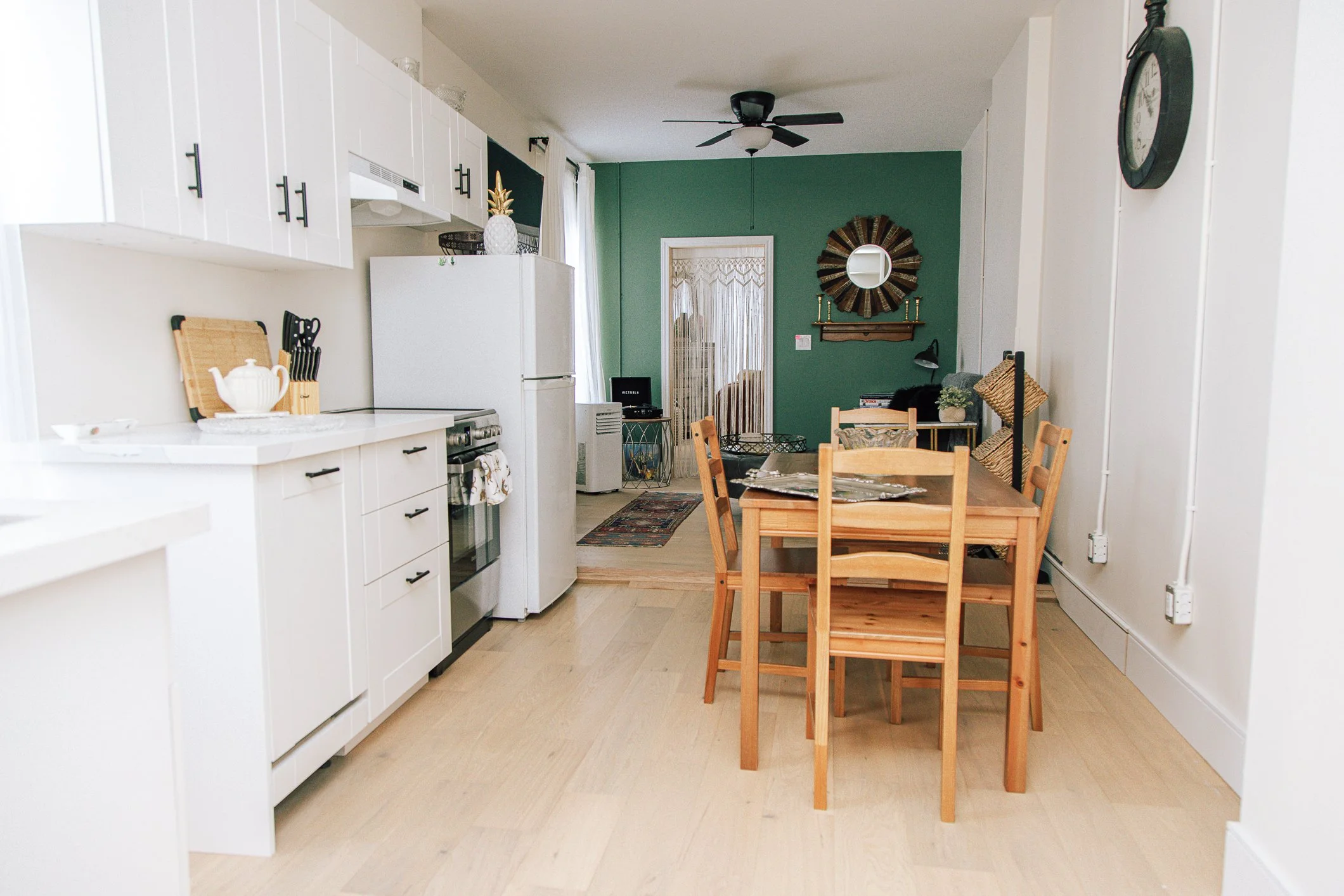 A cozy kitchen and dining area with white cabinets, a refrigerator, a wooden dining table with chairs, a green accent wall with a round mirror, and various decorative items.