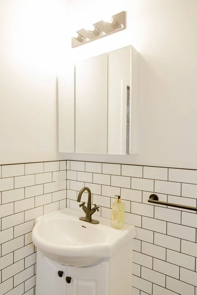 Small bathroom corner with white subway tile walls, white corner sink with a bronze faucet, a wall-mounted cabinet mirror above, a soap dispenser on the sink, and a towel bar on the wall.