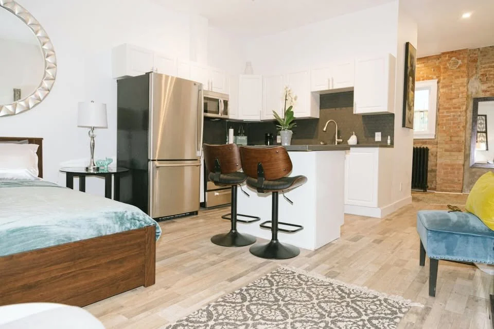 Open-concept living area with a bedroom, kitchen with white cabinets, stainless steel refrigerator, and a small seating area with two bar stools, a green plant on the counter, and a brick section of wall in the background.
