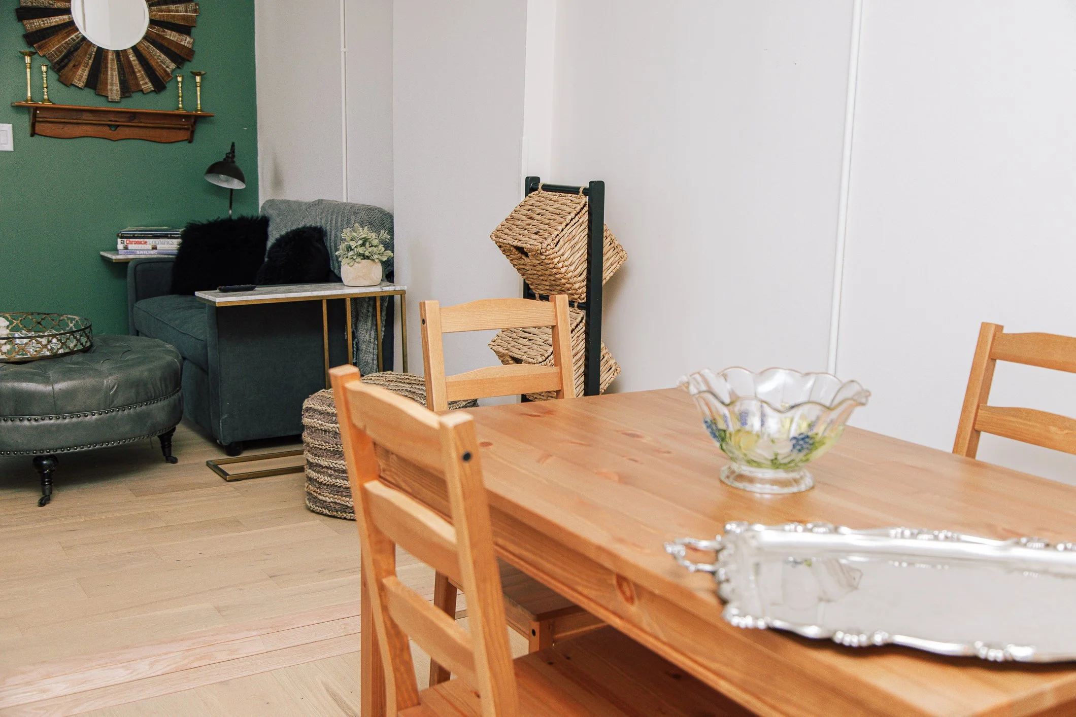 A dining room with a wooden table and chairs, a vase on the table, a gray couch with black pillows, a small bookshelf, a green wall with a round decorative mirror and candle holders.