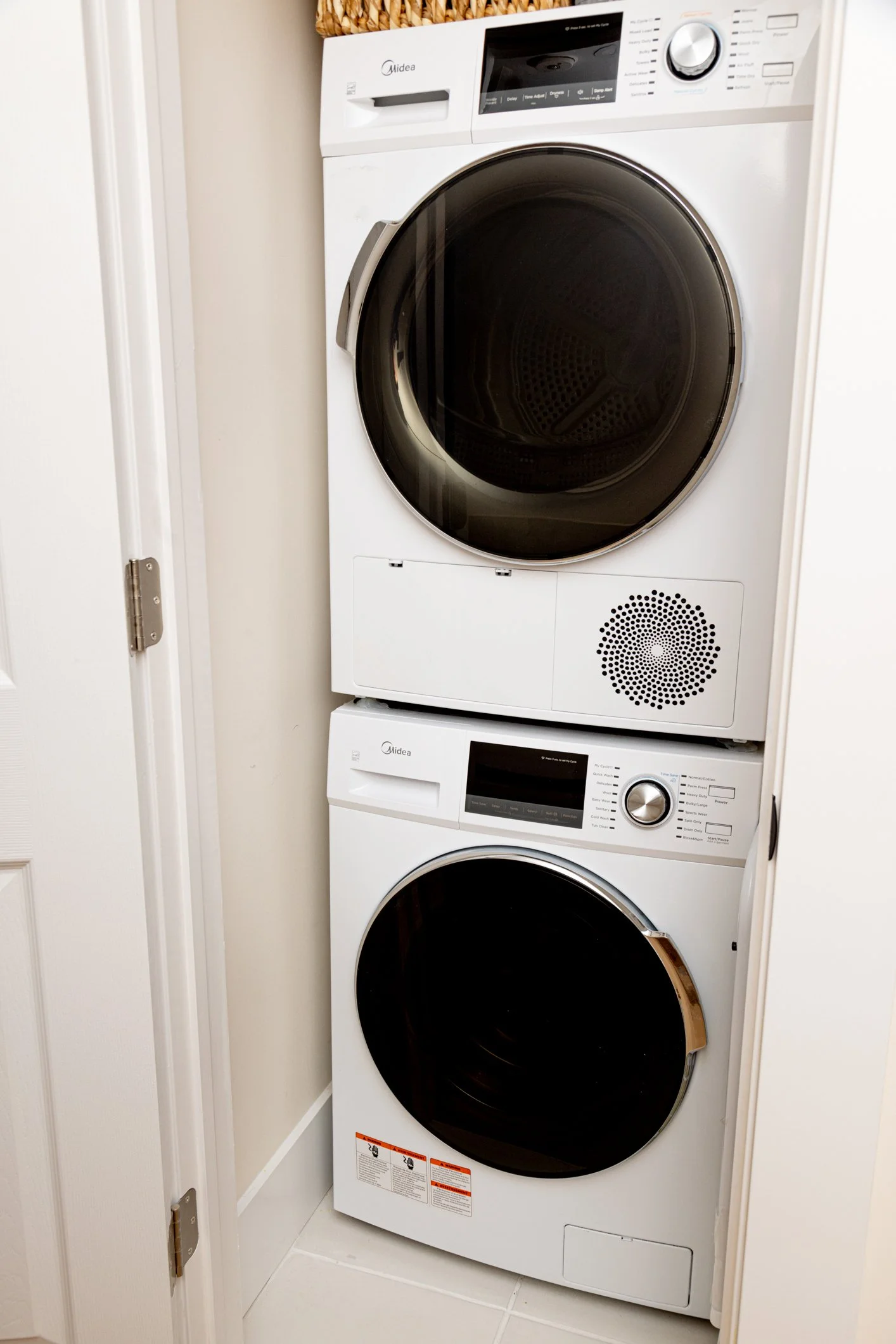 A stacked washer and dryer unit in a small laundry area. The machines are white with black doors; the top machine is a front-loading washing machine, and the bottom machine is a front-loading dryer.