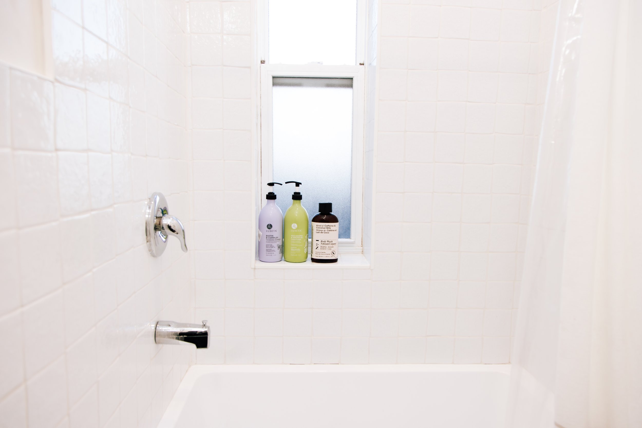 Close-up of a small bathroom window with three bottles of shower products on the windowsill, white tiled walls, and a chrome faucet.
