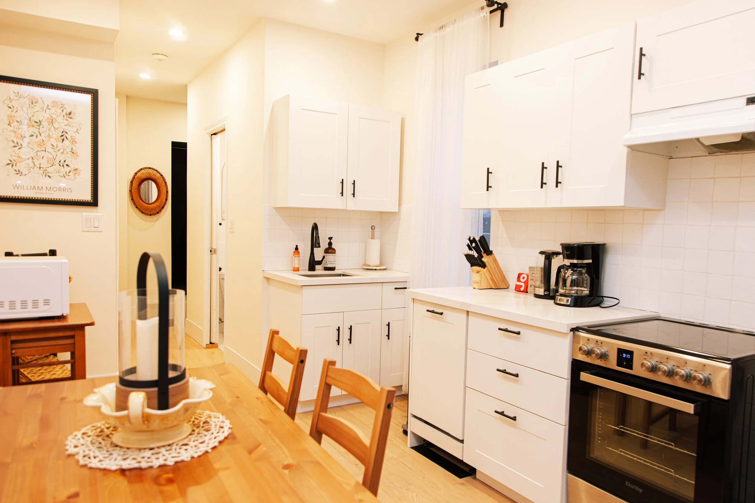 A cozy kitchen with white cabinets, a black stovetop, a coffee maker, a knife block, and a sink. There is a wooden dining table with chairs, a decorative cake stand with candles, wall art, and a window with white curtains.