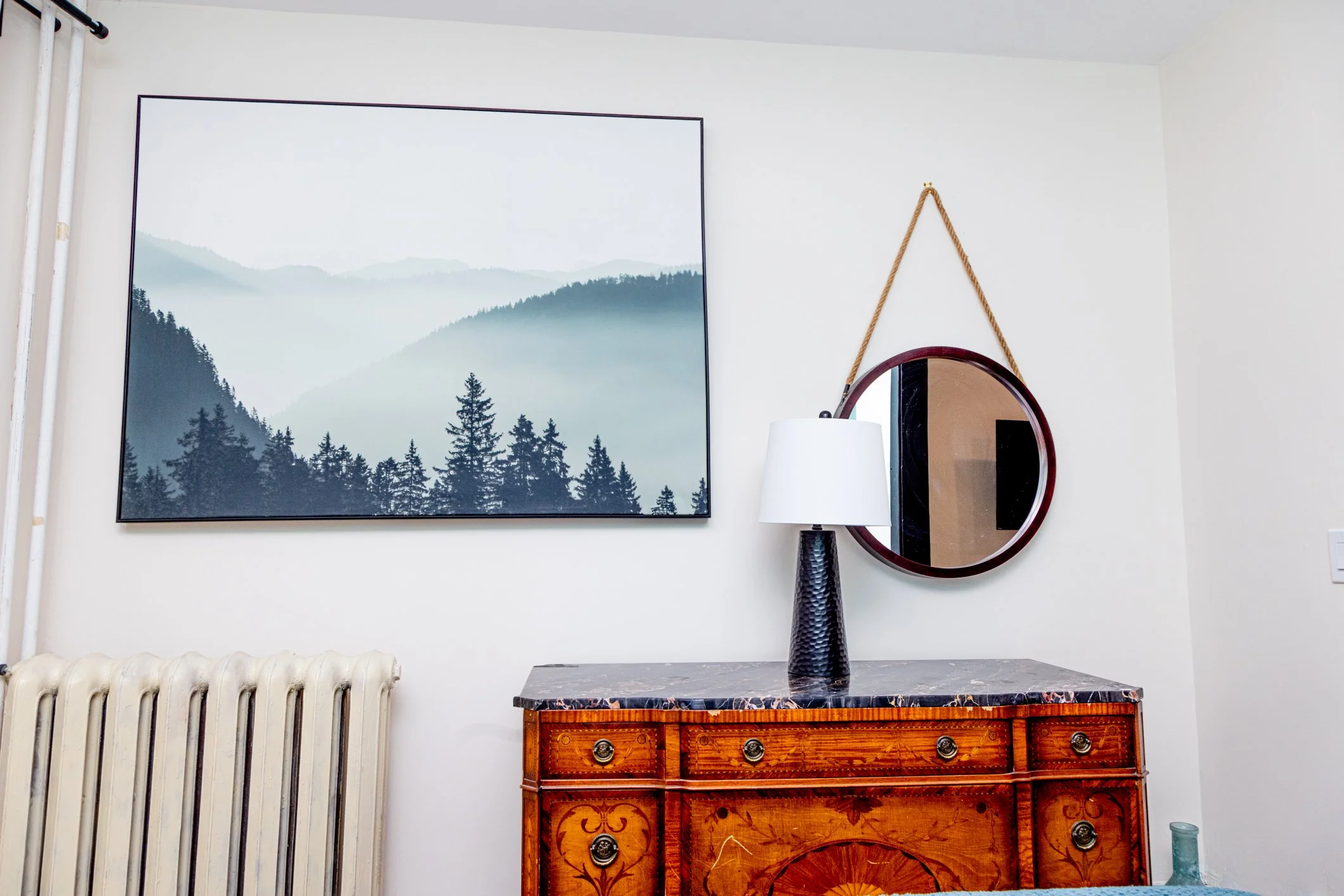 A room with a large wall art of mountains and trees, a white lamp on a dark stand, a round mirror hanging with a rope, a wooden dresser with ornate details, a white radiator, and a blue decorative item on the floor.