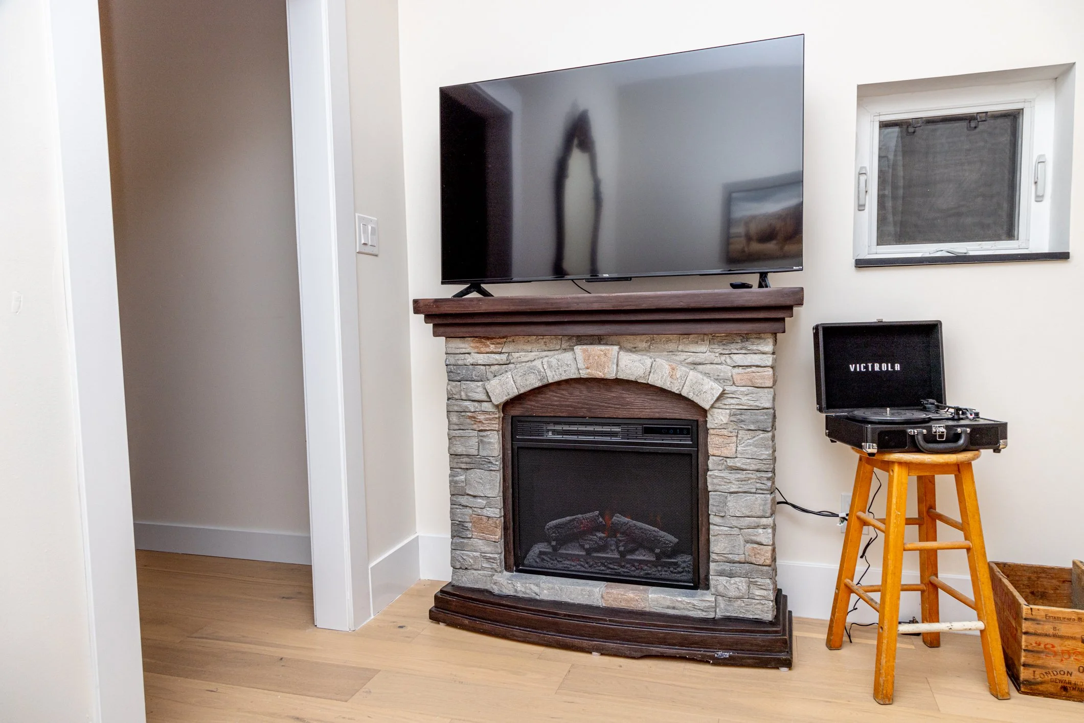 A living room corner with a TV mounted above a stone fireplace, a vinyl record player on a wooden stool, and a vintage wooden crate.