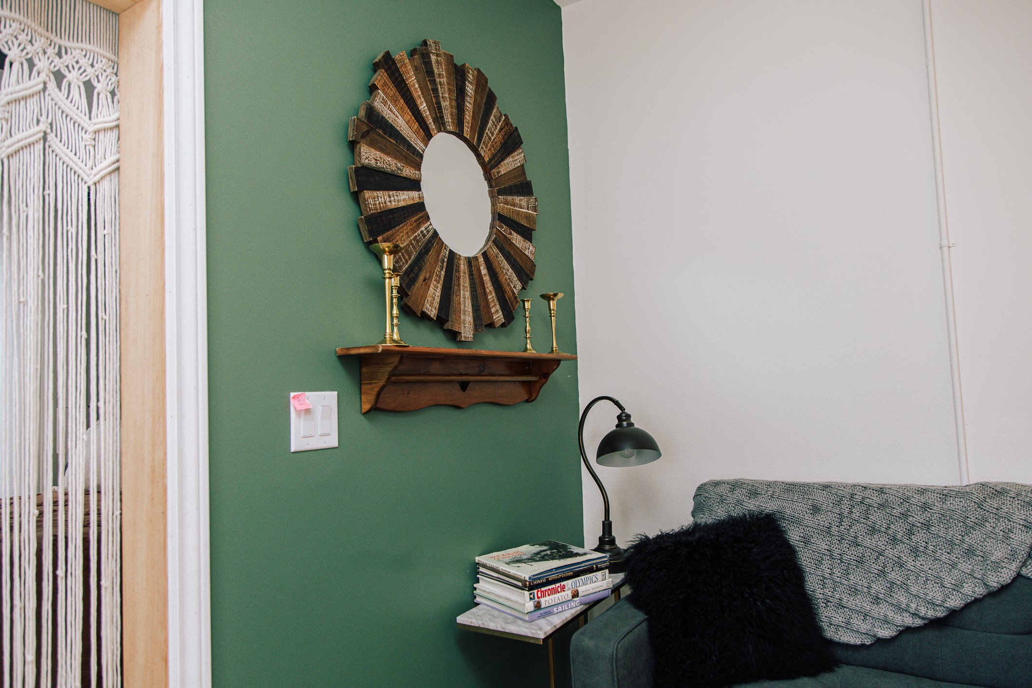 Interior corner of a living room with a green wall, a wooden shelf with brass candlesticks, a mirror with a wooden frame made of reclaimed wood, a stack of magazines, a black fur throw, and a desk lamp.