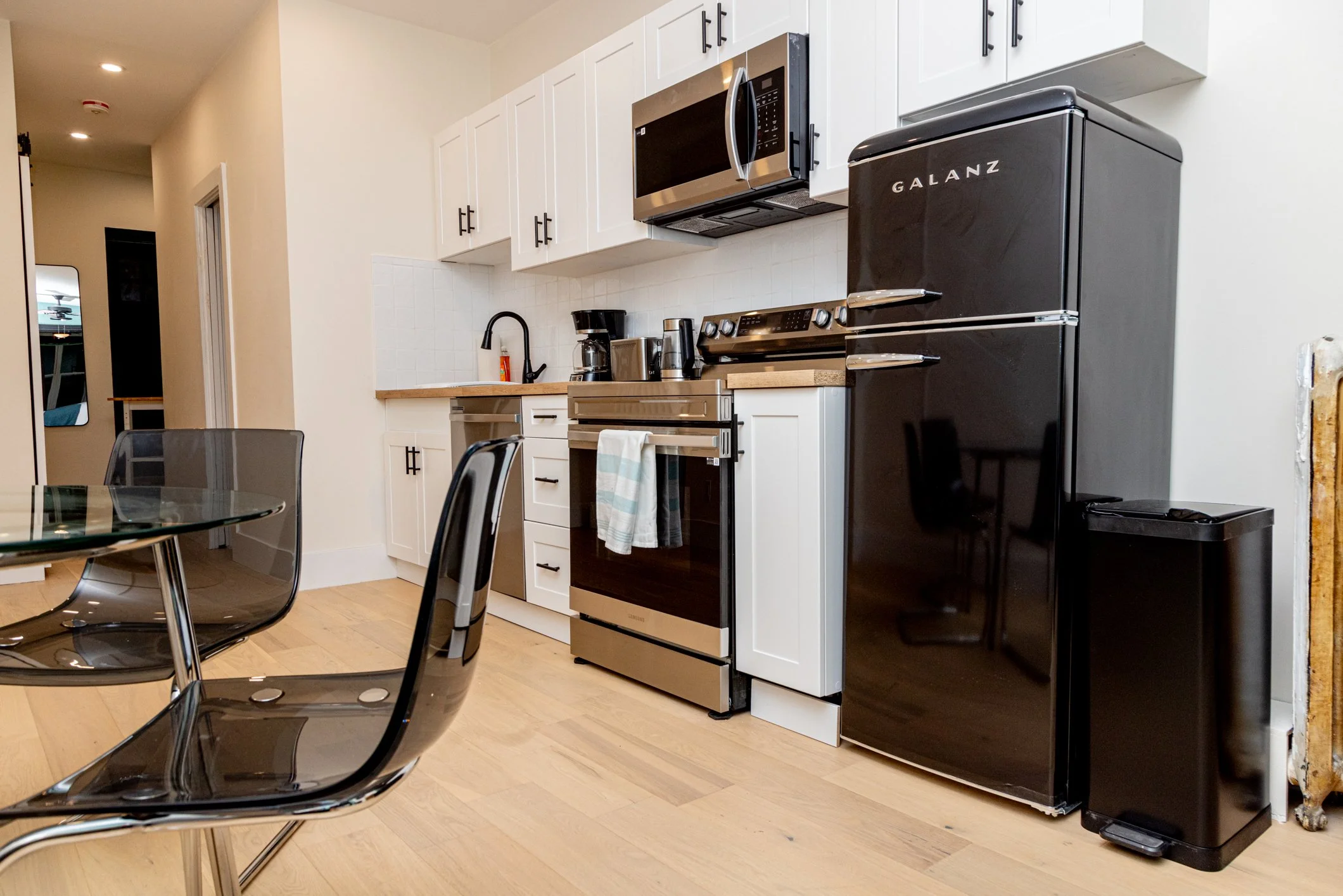 Modern kitchen with white upper cabinets, black handles, stainless steel microwave, black Galanz refrigerator, coffee maker, and hardwood flooring. Part of a dining table with black chairs is visible in the foreground.