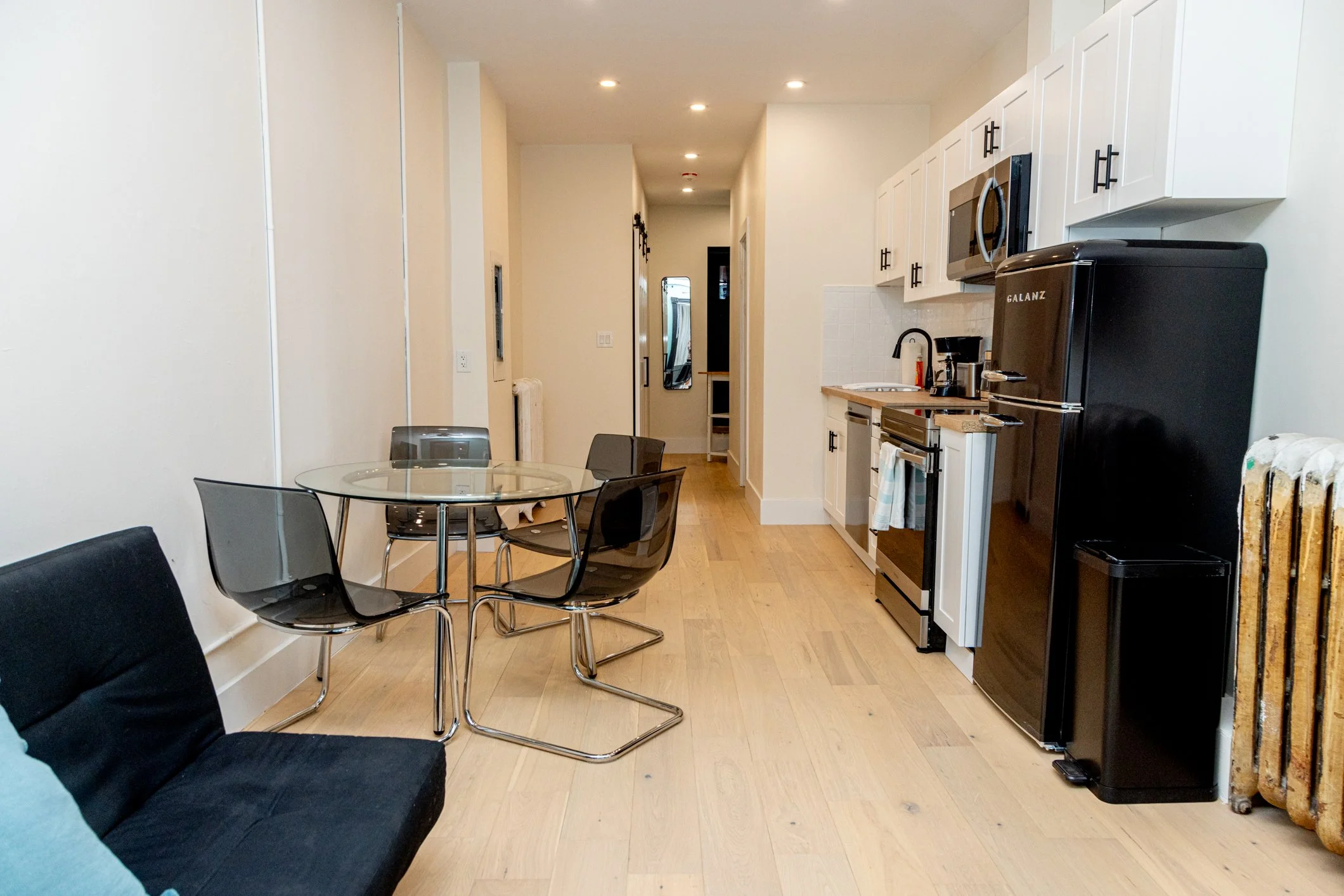 Interior view of a modern kitchen and dining area with white cabinets, black refrigerator, microwave, coffee maker, glass dining table, and black chairs. Hardwood floors and recessed ceiling lights.