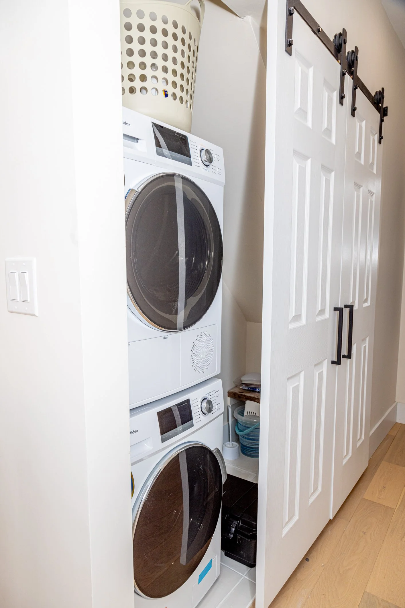 A small laundry closet with a stacked washer and dryer, a white cabinet with black handles, a basket on top of the washer, and cleaning supplies below.