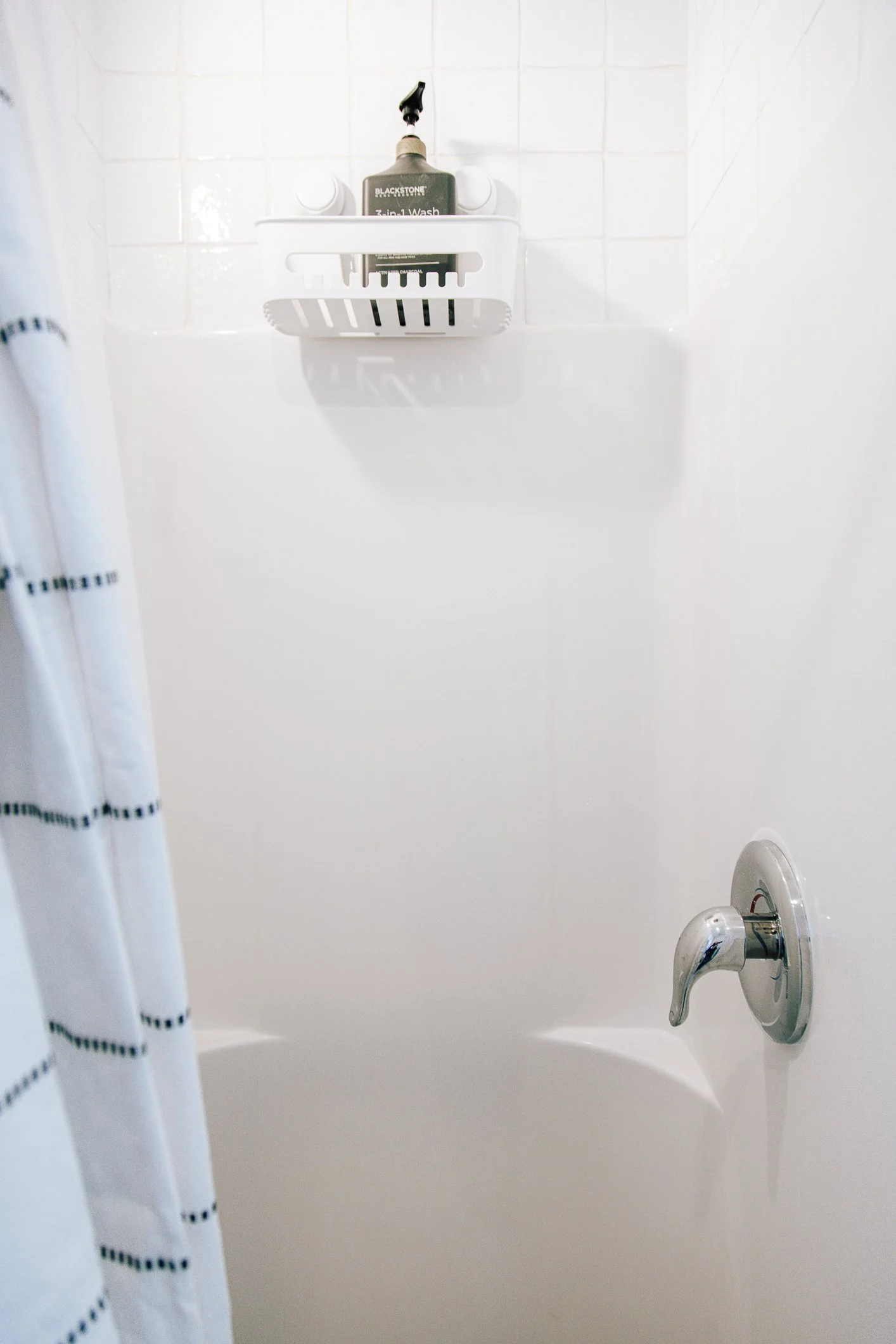 Bathroom shower with white tiled wall, a soap dish holding a bottle of shower gel, a curtain with black stripes, and a silver bathroom faucet.