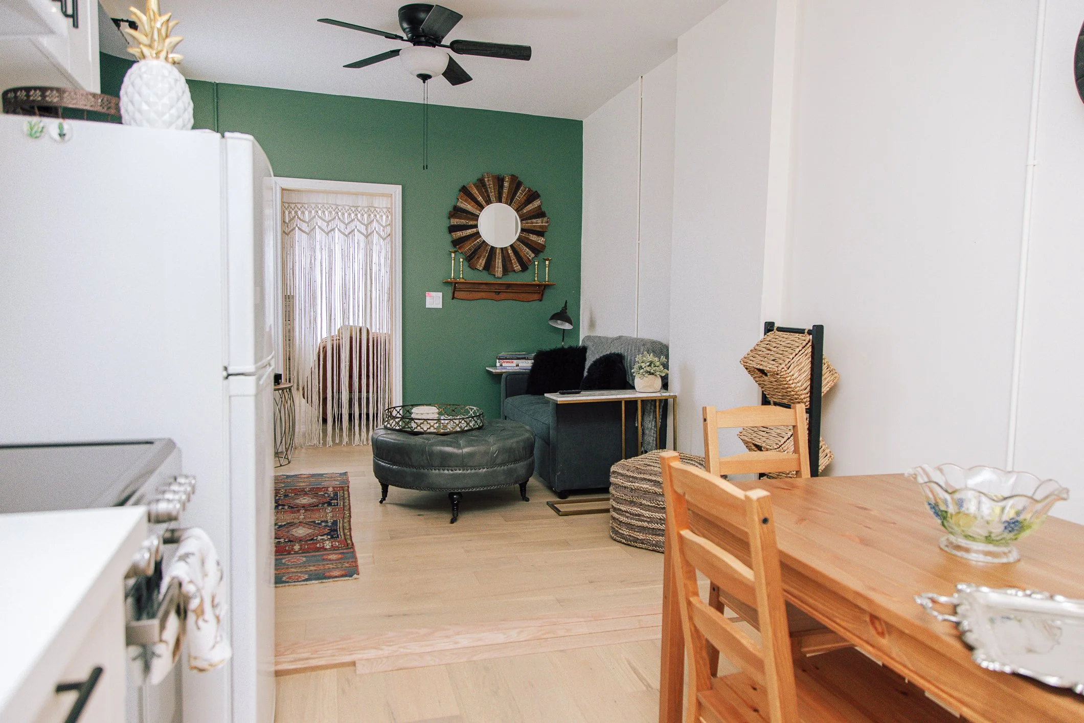 Interior view of a living area with a green accent wall, a ceiling fan, a white beaded curtain, a gray armchair, a small side table with a plant, and various decorative items including a large circular mirror and candlesticks. A wooden dining table w