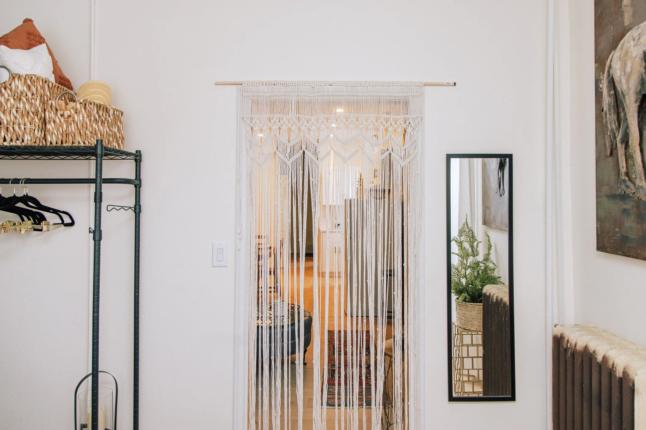 Interior of a cozy living space with bohemian decor, including a beaded curtain doorway, a mirror, potted plants, a wall painting, and a small blurred living room visible through the curtain.