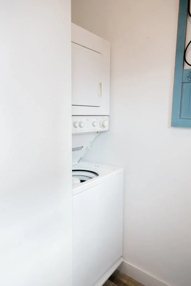 A white stacked washer and dryer unit in a small laundry space with white walls and a wooden floor.