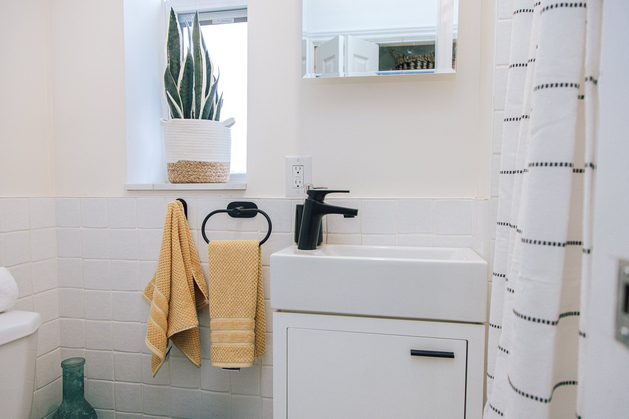 A small bathroom with a white sink, a black faucet, a large potted snake plant on the windowsill, a mirror above the sink, and two yellow towels hanging on a black towel ring. There's a white laundry machine to the right of the sink, a small blue vas
