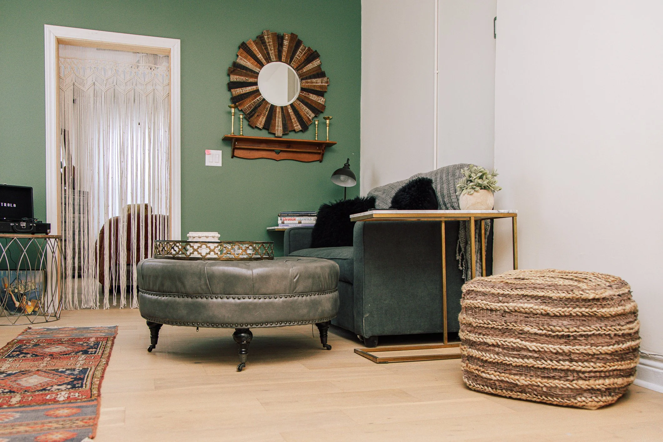 Living room with green and white walls, a gray sofa with black fur pillows, a round beige ottoman with a tray on top, a wooden table with woven baskets, a mirror with a sunburst frame, a beaded curtain door, and a small side table with books and a la