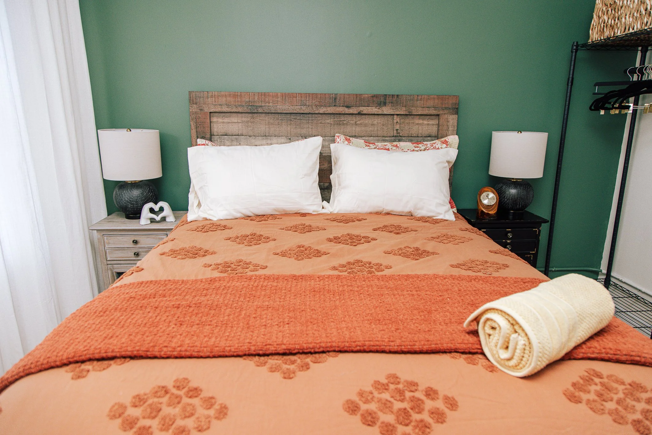 A neatly made bed with an orange bedspread decorated with floral patterns, two rolled towels at the foot, white pillows with floral pillowcases, a rustic wooden headboard, side tables with lamps, and a clothing rack in a room with green walls.