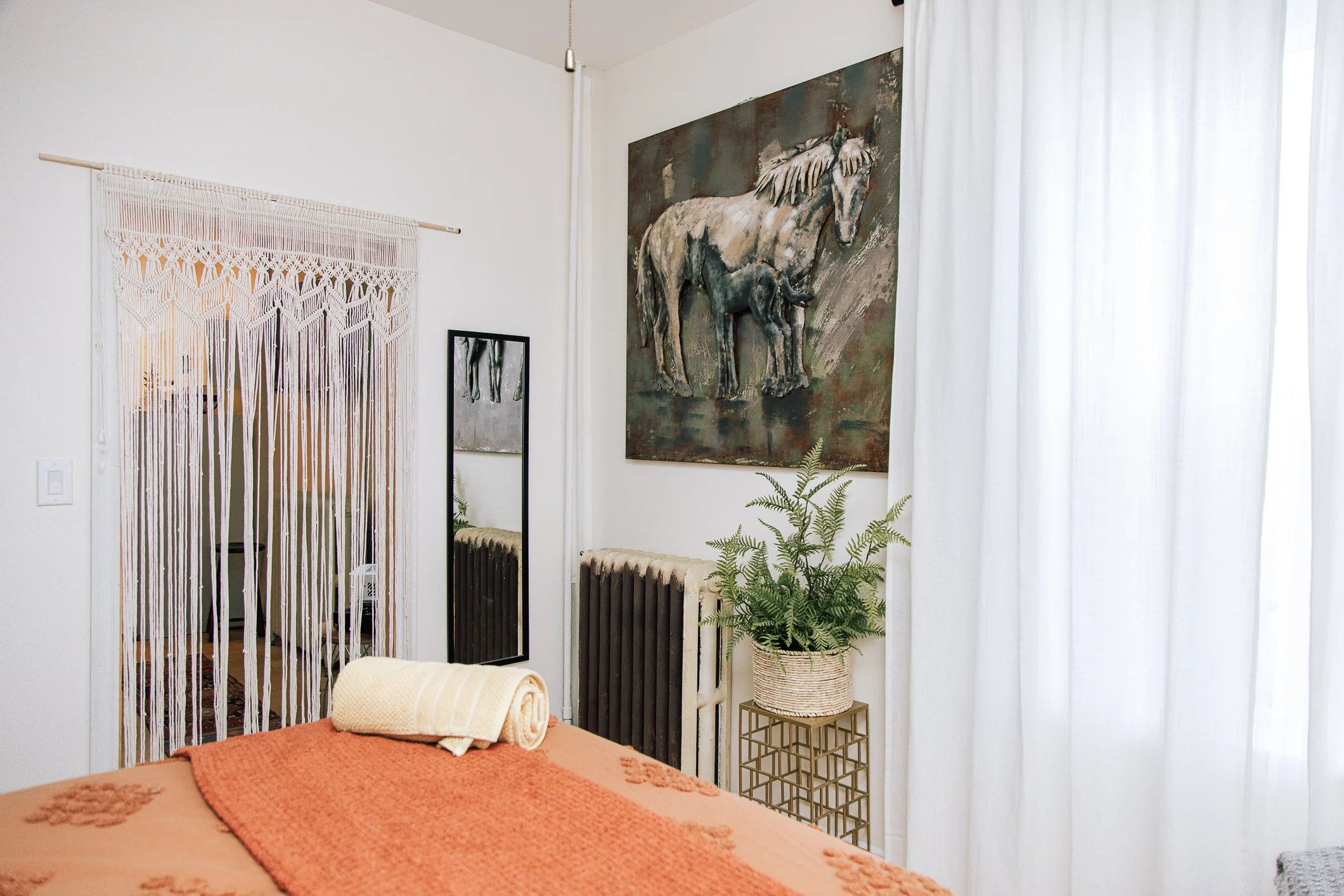 Interior of a bedroom with a bed covered in an orange blanket, a rolled white towel, and a smaller towel on top. The wall has a large painting of a white horse and a smaller framed artwork. There is a metal side table with a potted fern and a radiato