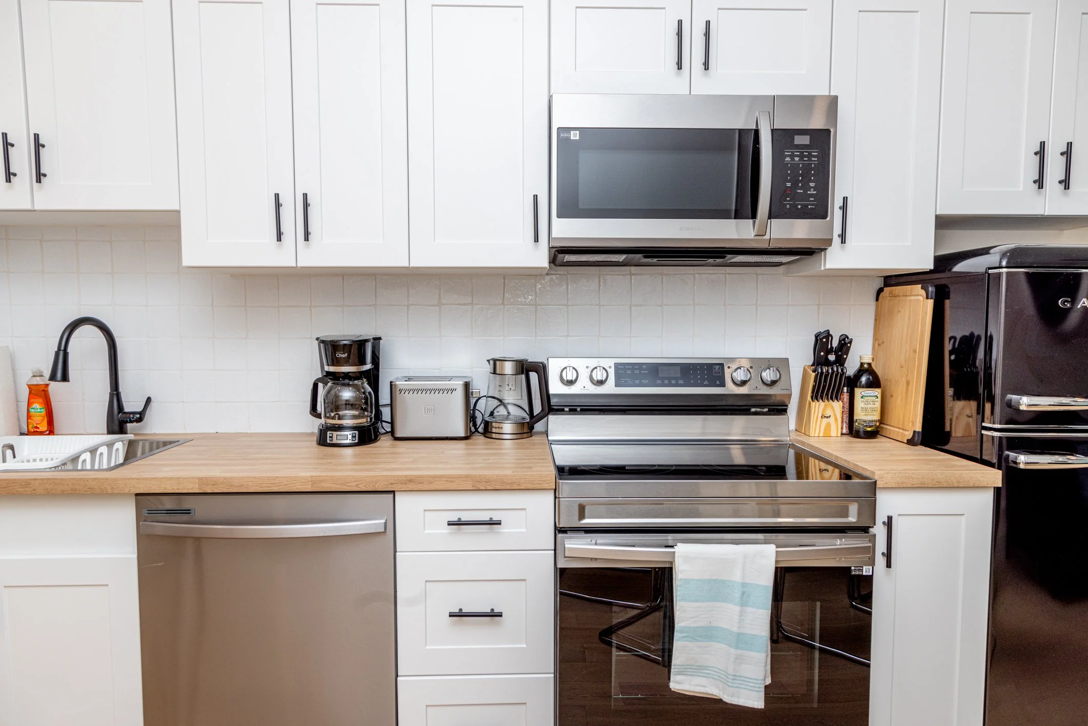 Modern kitchen with white cabinets, stainless steel microwave and stove, coffee maker, toaster, and black appliances on a wooden countertop.