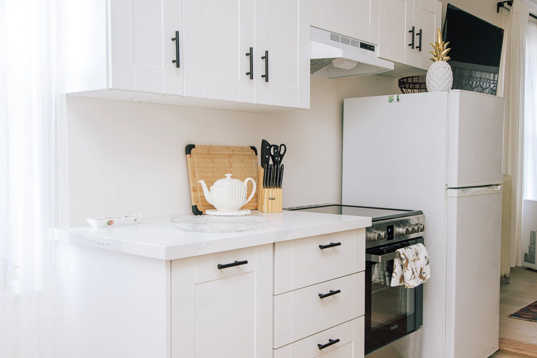 A kitchen with white cabinets, a white countertop with a teapot on a carved glass tray, a wooden cutting board, and a knife block. There is a stove with a towel hanging from the handle, and a white refrigerator. On top of the refrigerator, there is a
