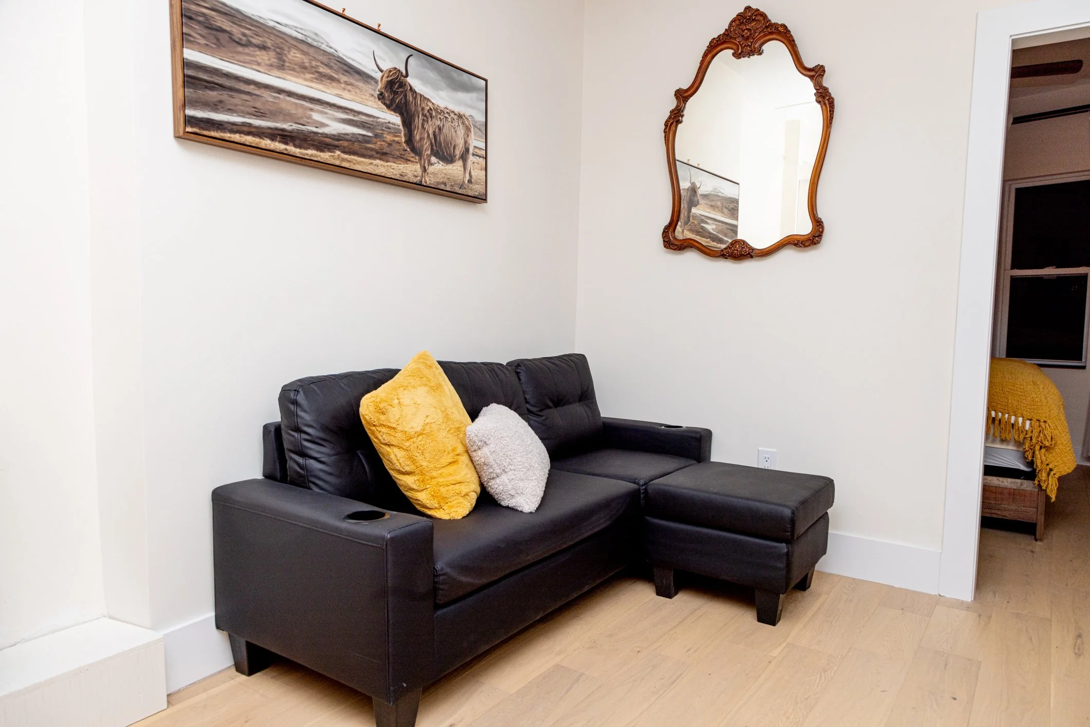 A black leather sofa with three decorative pillows, one yellow, one gray, and one white, in a room with hardwood floors, a framed painting of a highland cow, and a vintage mirror on the wall.