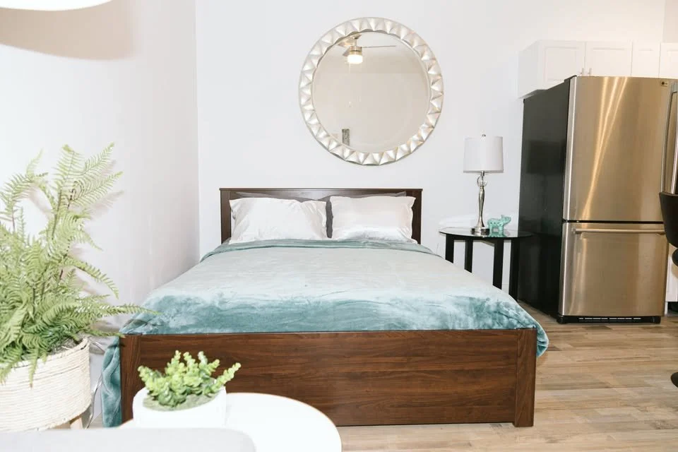 A minimalist bedroom with a wooden bed frame, white bedding, and a blue blanket. A round mirror hangs above the bed. To the right, there is a black side table with a modern lamp and decorative item. A stainless steel refrigerator stands next to the t