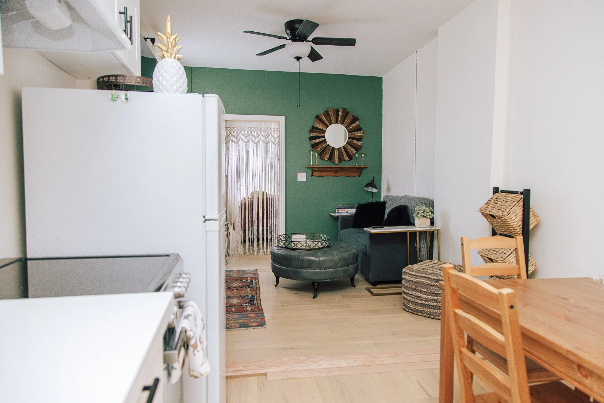 Interior of a living room with a green accent wall, a black ceiling fan, a round mirror with a decorative frame, a wooden shelf with candlesticks, a small side table with magazines and a lamp, a gray couch with pillows, a woven basket, and part of a 