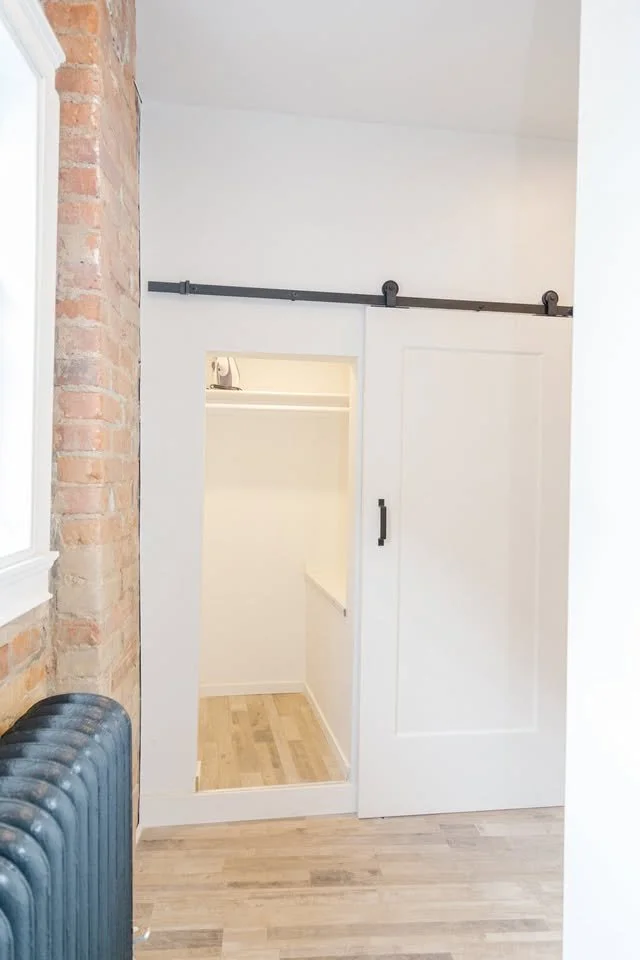 A small walk-in closet with white walls, a black sliding barn door, a black handle, and a sliding track. Part of an exposed brick wall and a radiator are visible in the room.