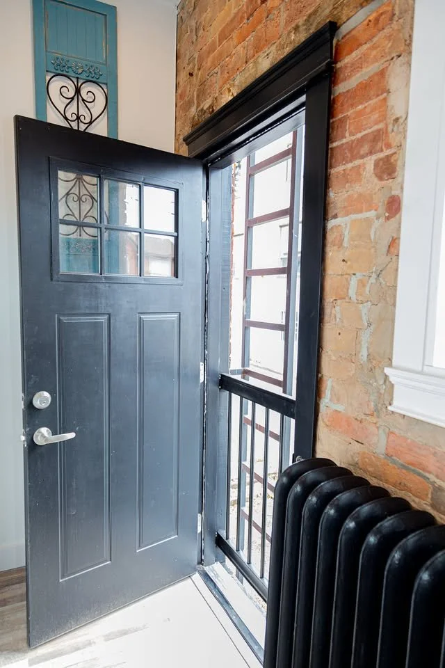Open front door with a small railing, brick wall, radiator, and window.