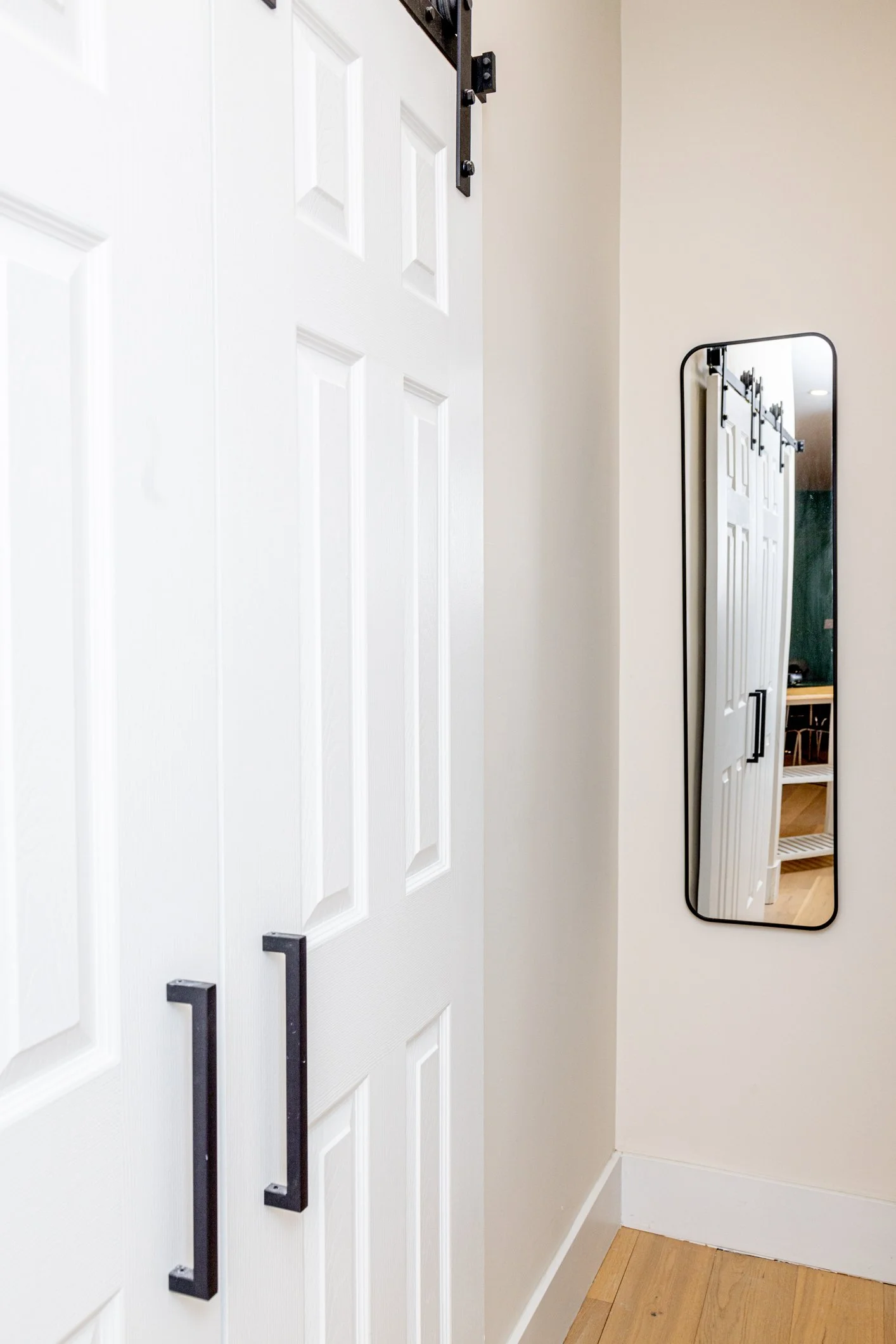 Interior of a home with a white door, black hardware, a tall vertical mirror on the wall, and hardwood flooring.