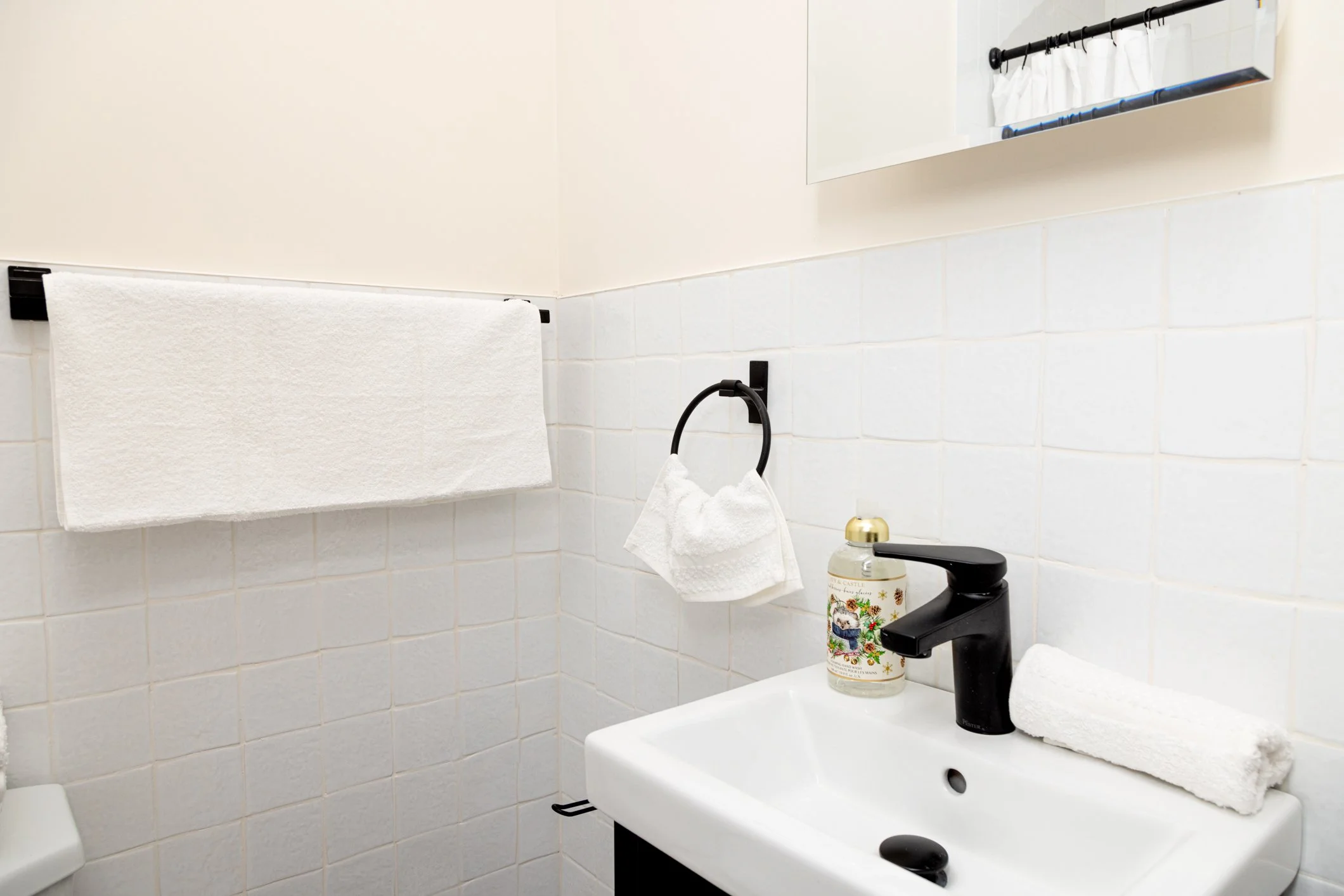 Bathroom sink with black faucet, hand towel on sponge holder, decorative soap dispenser, mirror, curtain over window, towel rail.
