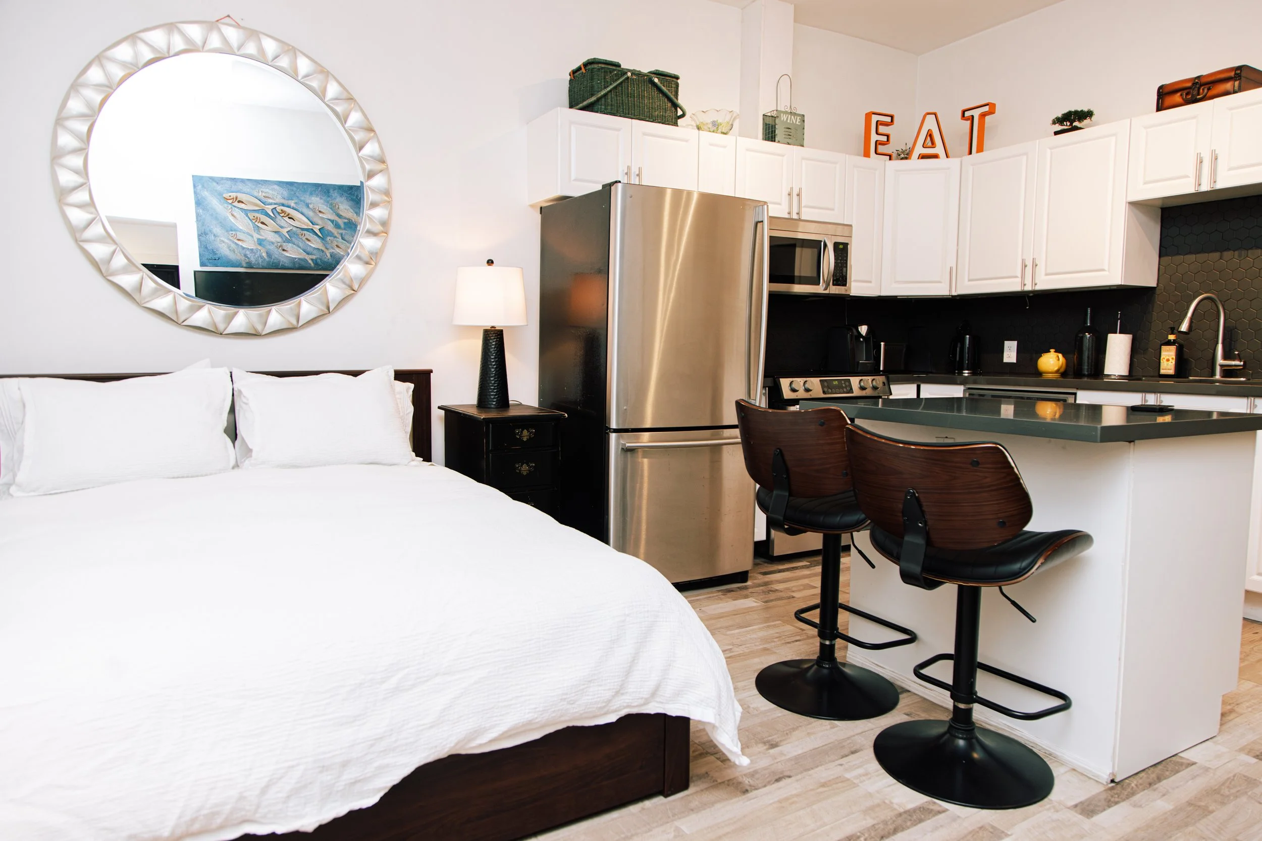 A studio apartment showing a bed with white bedding, a circular mirror on the wall, a nightstand with a lamp, a refrigerator, a microwave, and a kitchen area with white cabinets, black backsplash, and a small dining table with two black chairs.
