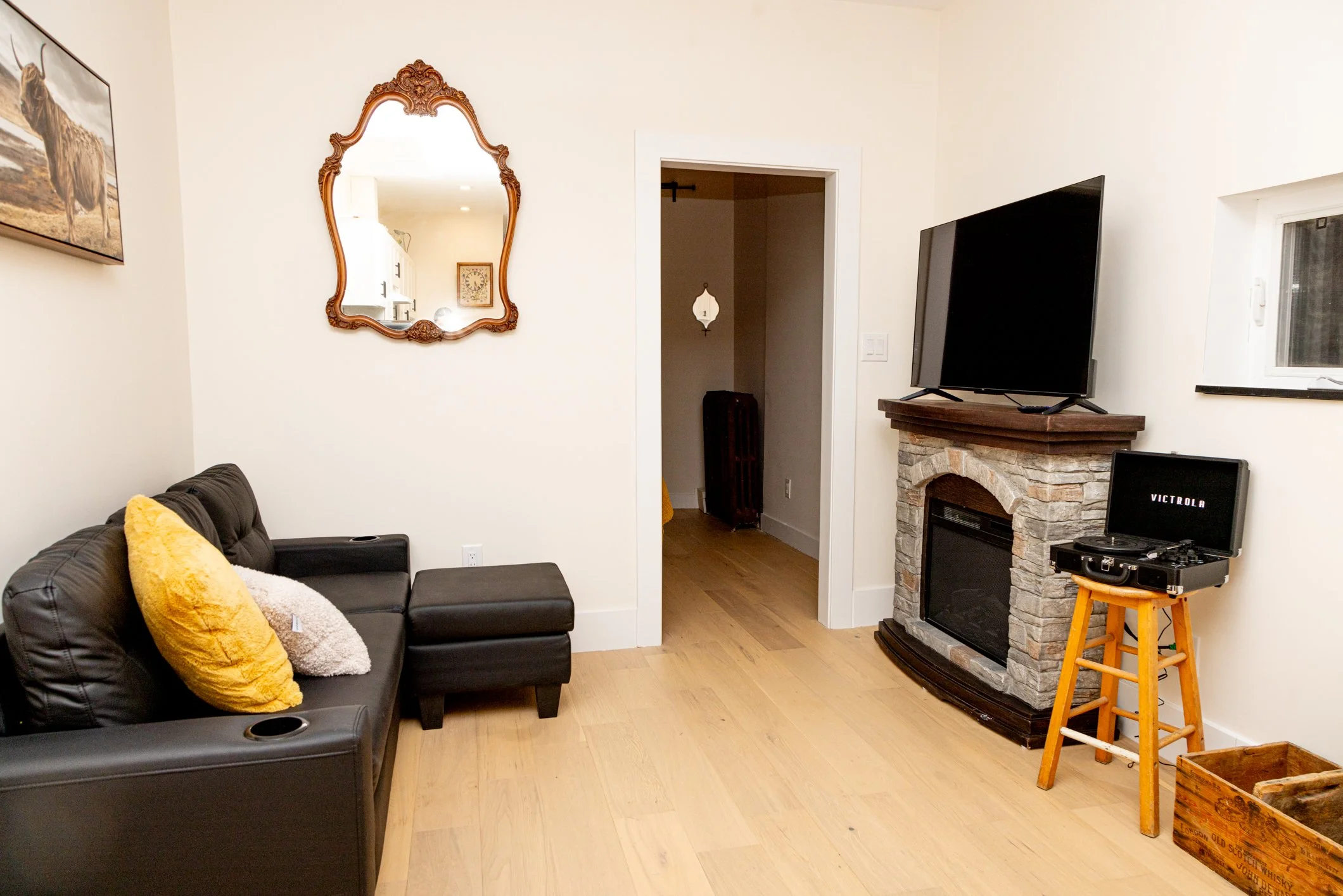 A cozy living room with a black leather couch decorated with yellow and beige throw pillows, a wall-mounted mirror with an ornate wooden frame, a fireplace with a stone surround, a flat-screen TV, a wooden stool with a turntable, a window, and wooden