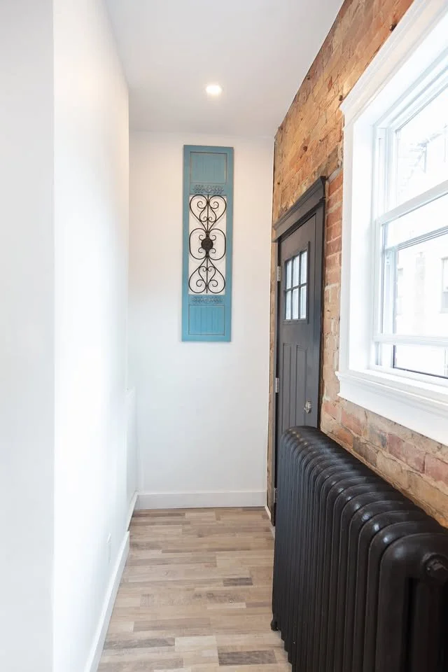 A narrow hallway with a white wall on the left, a brick wall with a window on the right, and a black radiator along the brick wall. There's a small dark door with window panes on the door, and a decorative wall hanging on a blue frame at the end of t