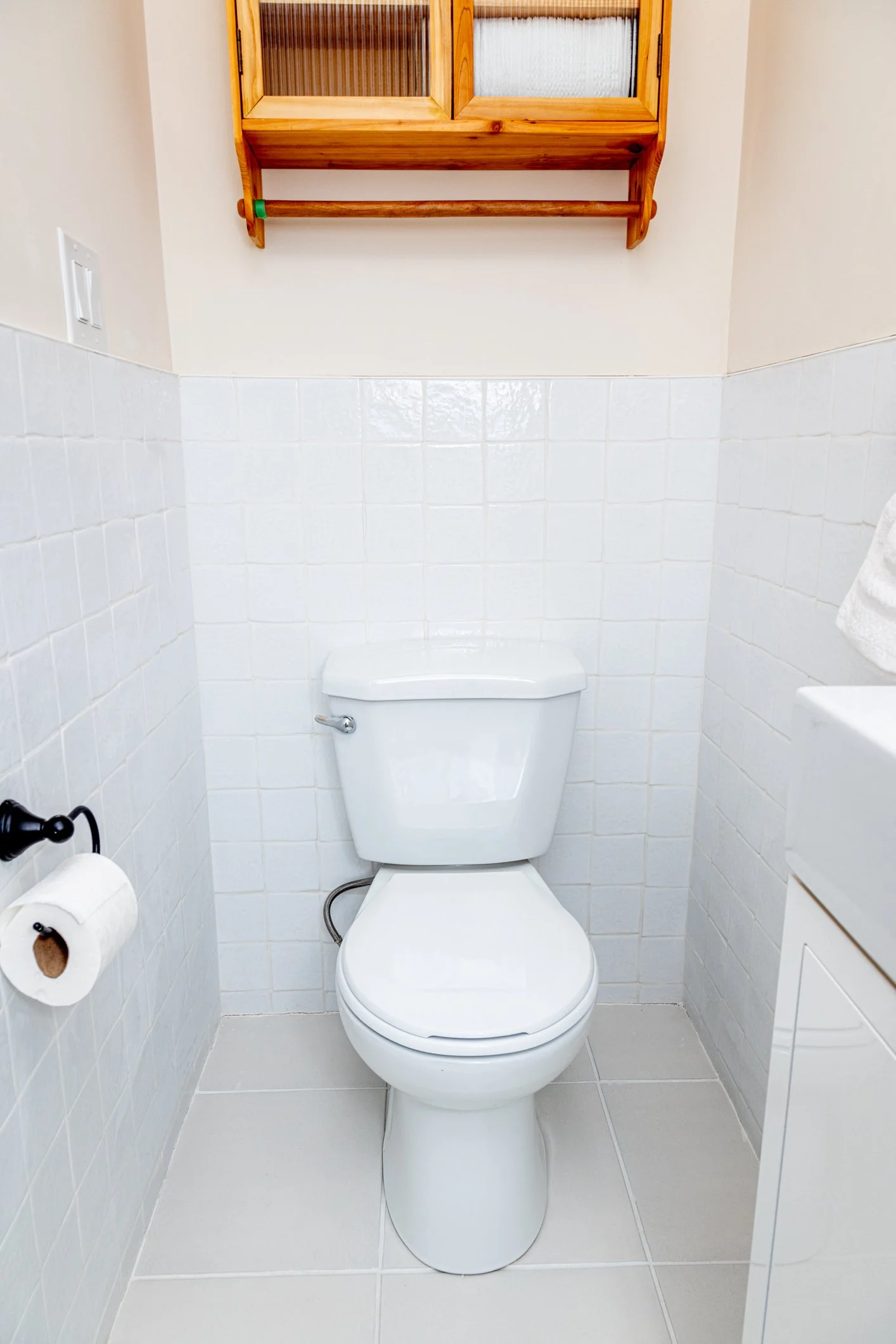 A small bathroom with white tiled walls and a white toilet. On the left wall, there is a toilet paper roll. Above the toilet, a wooden cabinet with glass doors is mounted on the wall.