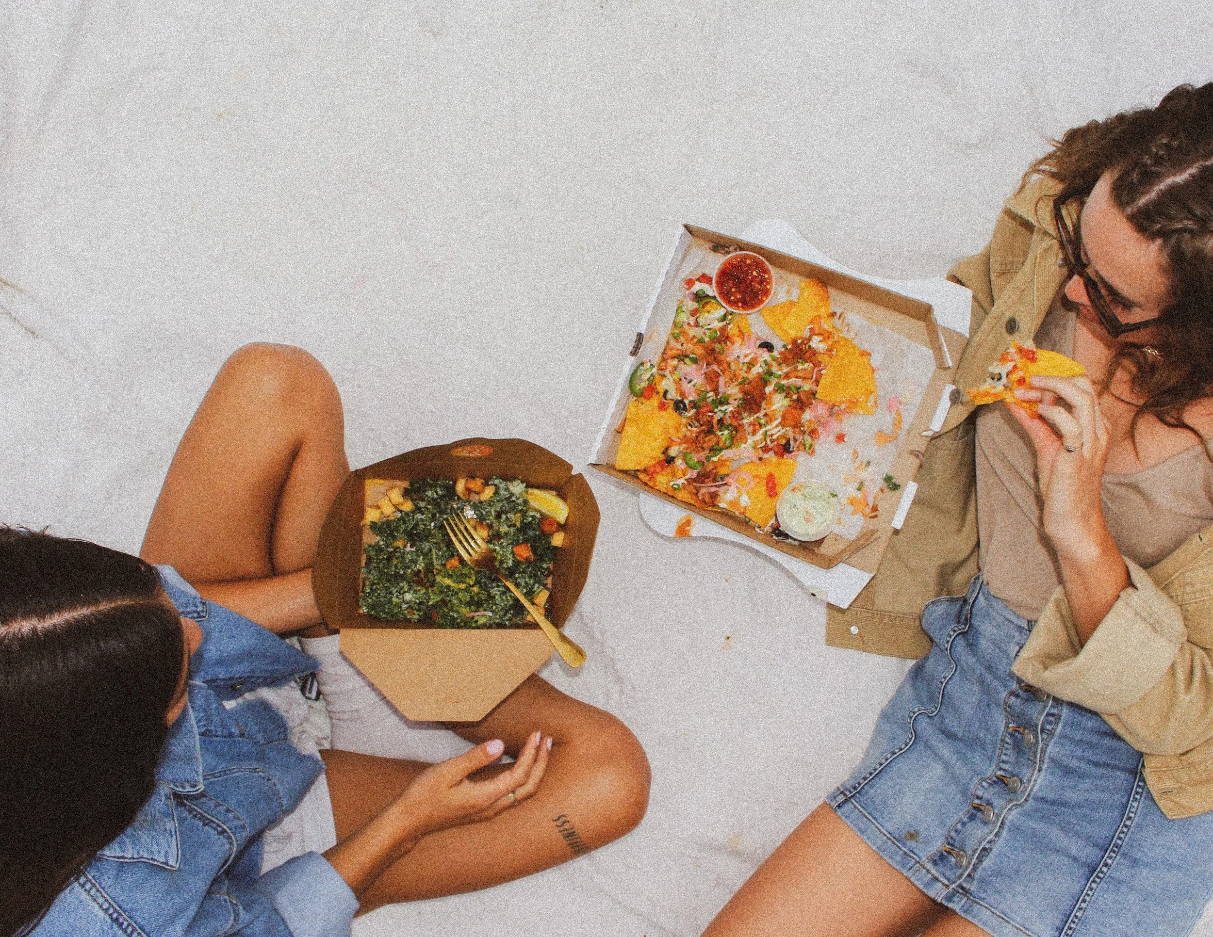 Two women sitting on a white floor eating food, with a salad in a brown bowl. Restaurant photography for vegan market