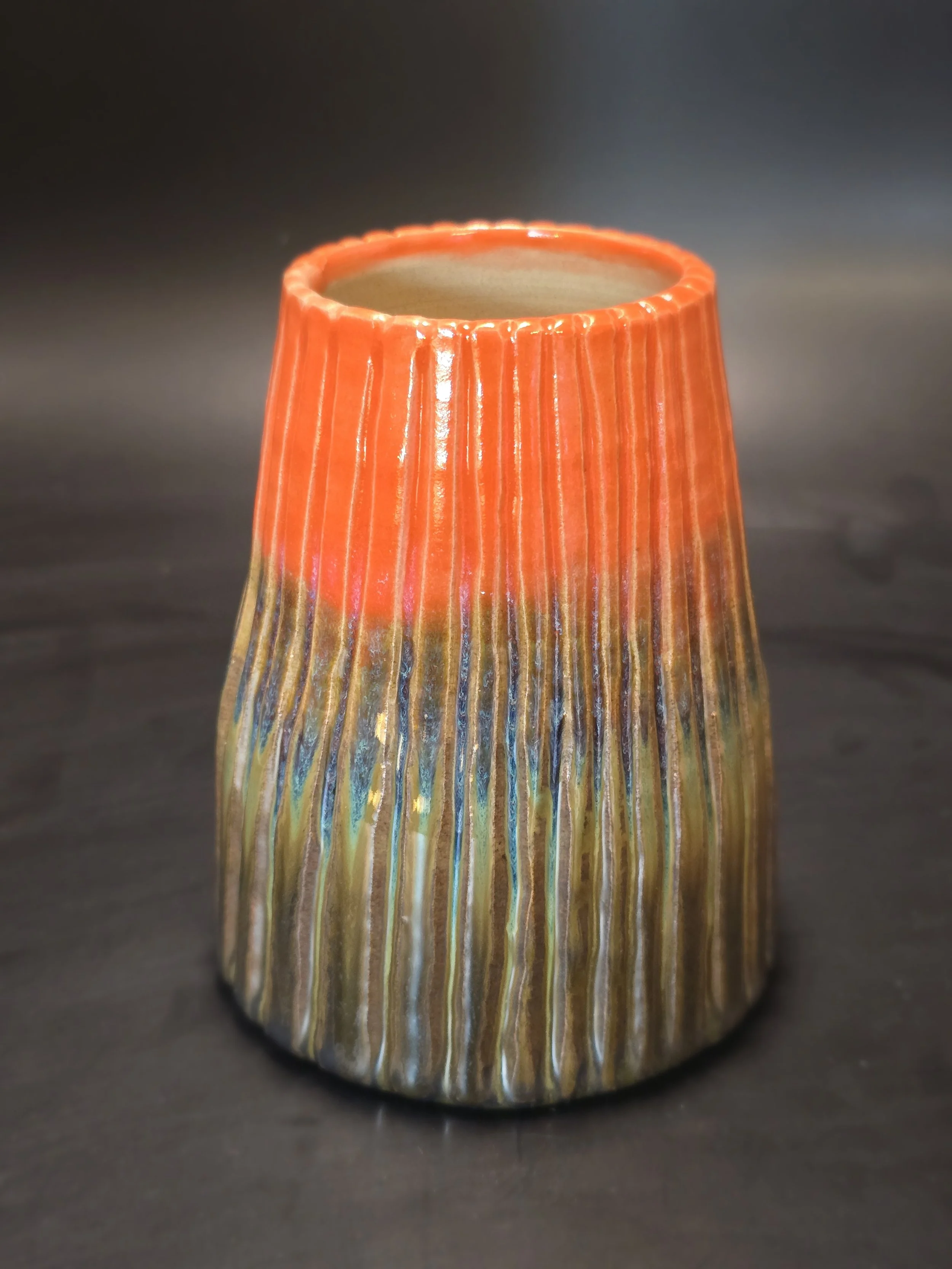 Colorful ceramic vase with an orange top and earthy tones at the base, placed on a dark surface.