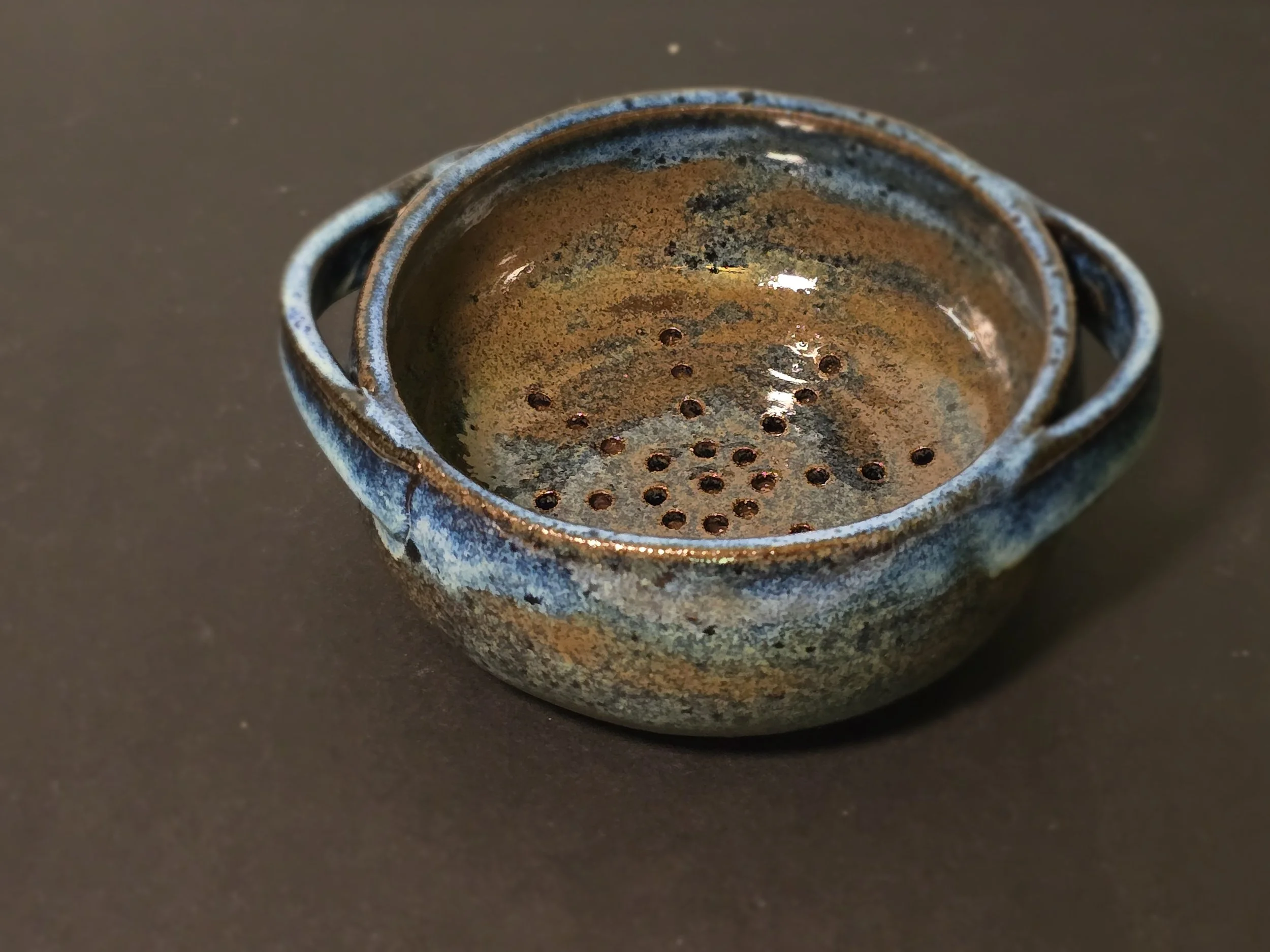 A small handmade ceramic tea strainer with a rustic glazed finish and two small handles, sitting on a dark surface.