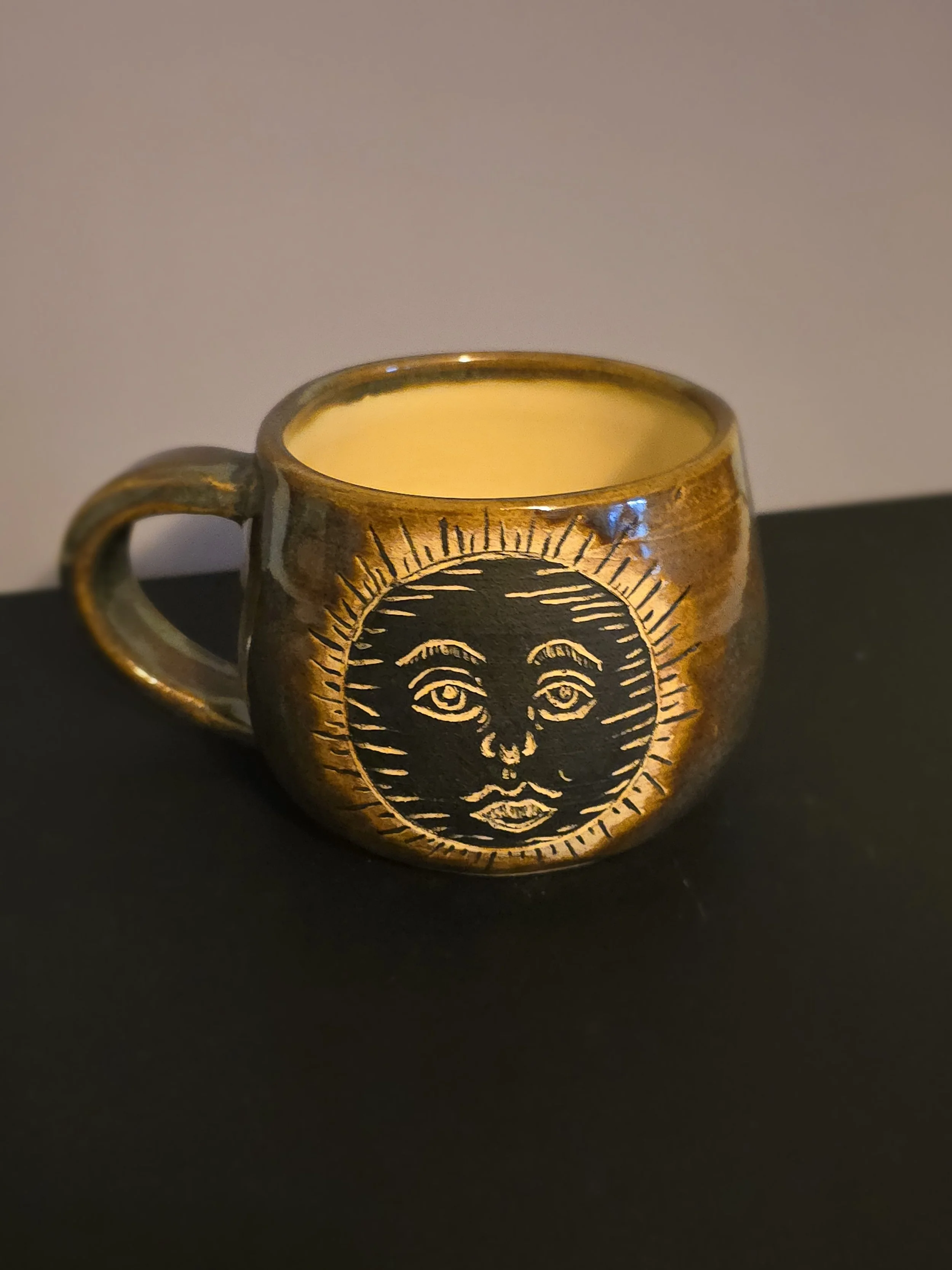 Ceramic coffee mug with a black sun face design on a gold background, placed on a black surface.