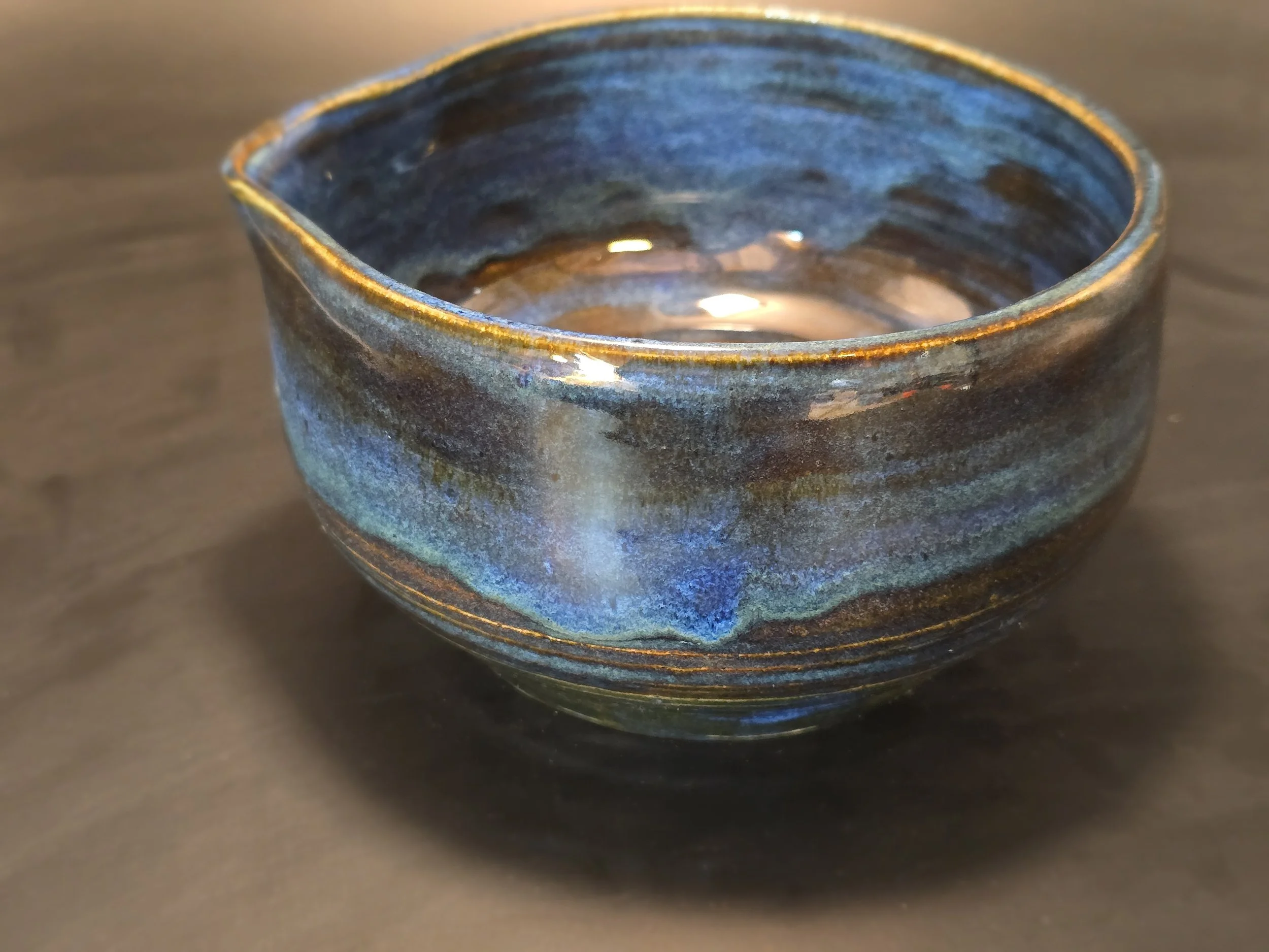 A round ceramic bowl with a glossy blue and brown glaze, empty and placed on a dark surface.