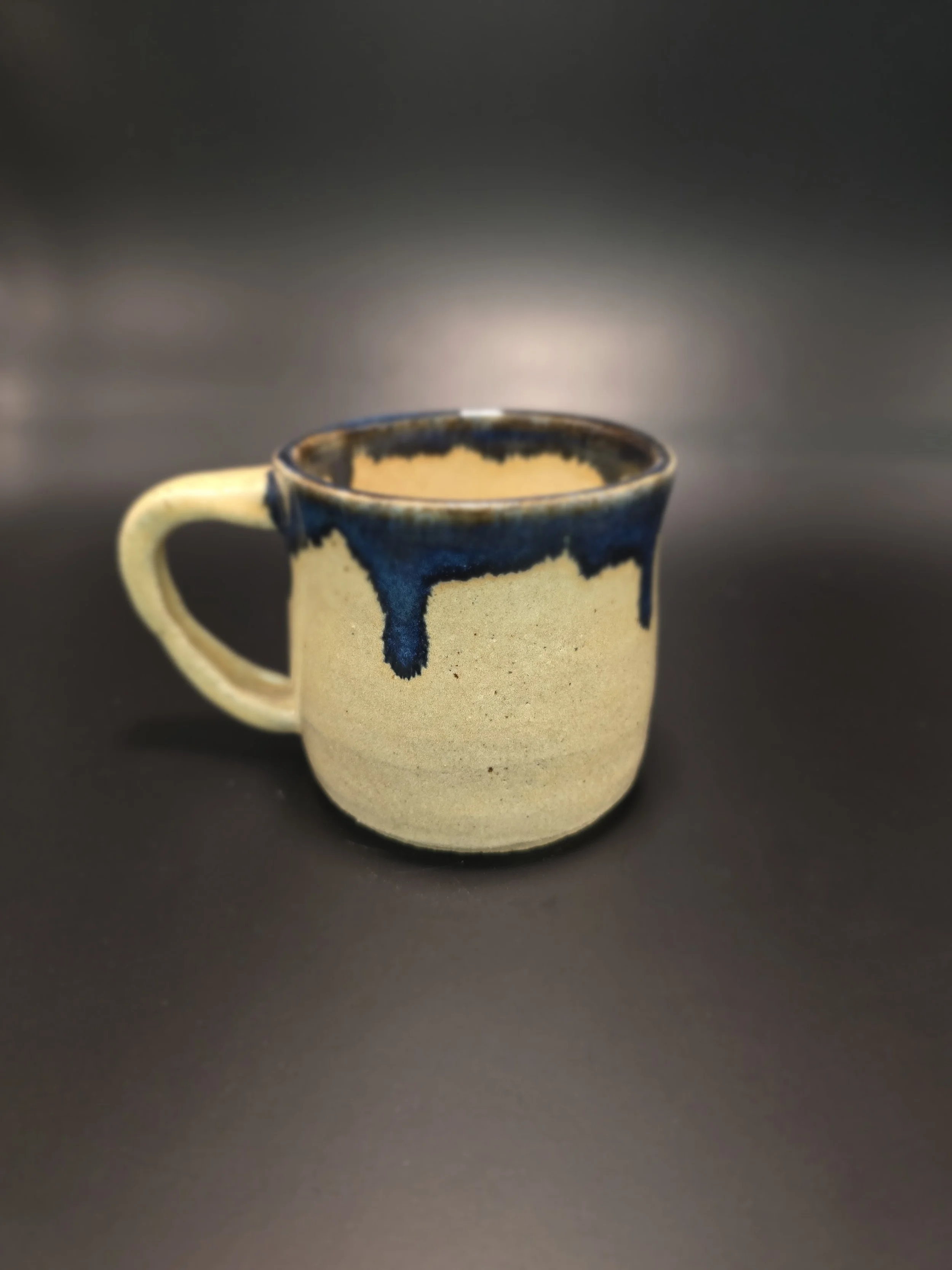 A beige ceramic mug with a dark blue dripping glaze around the rim and down the sides, placed on a black surface with a dark background.