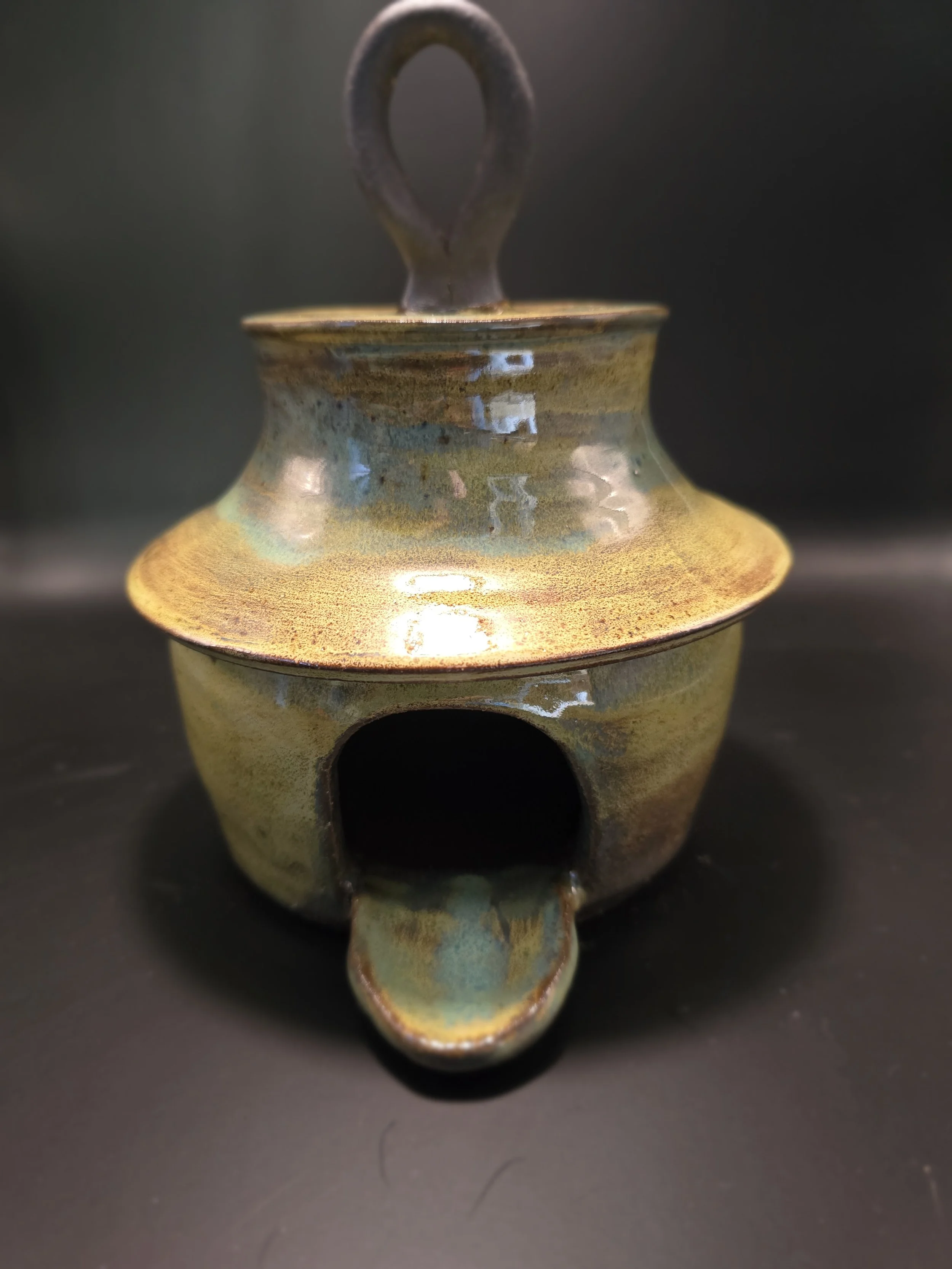 A ceramic oil burner with a round opening at the front and a small handle on the top, glazed with a mixed earthy-toned glaze.