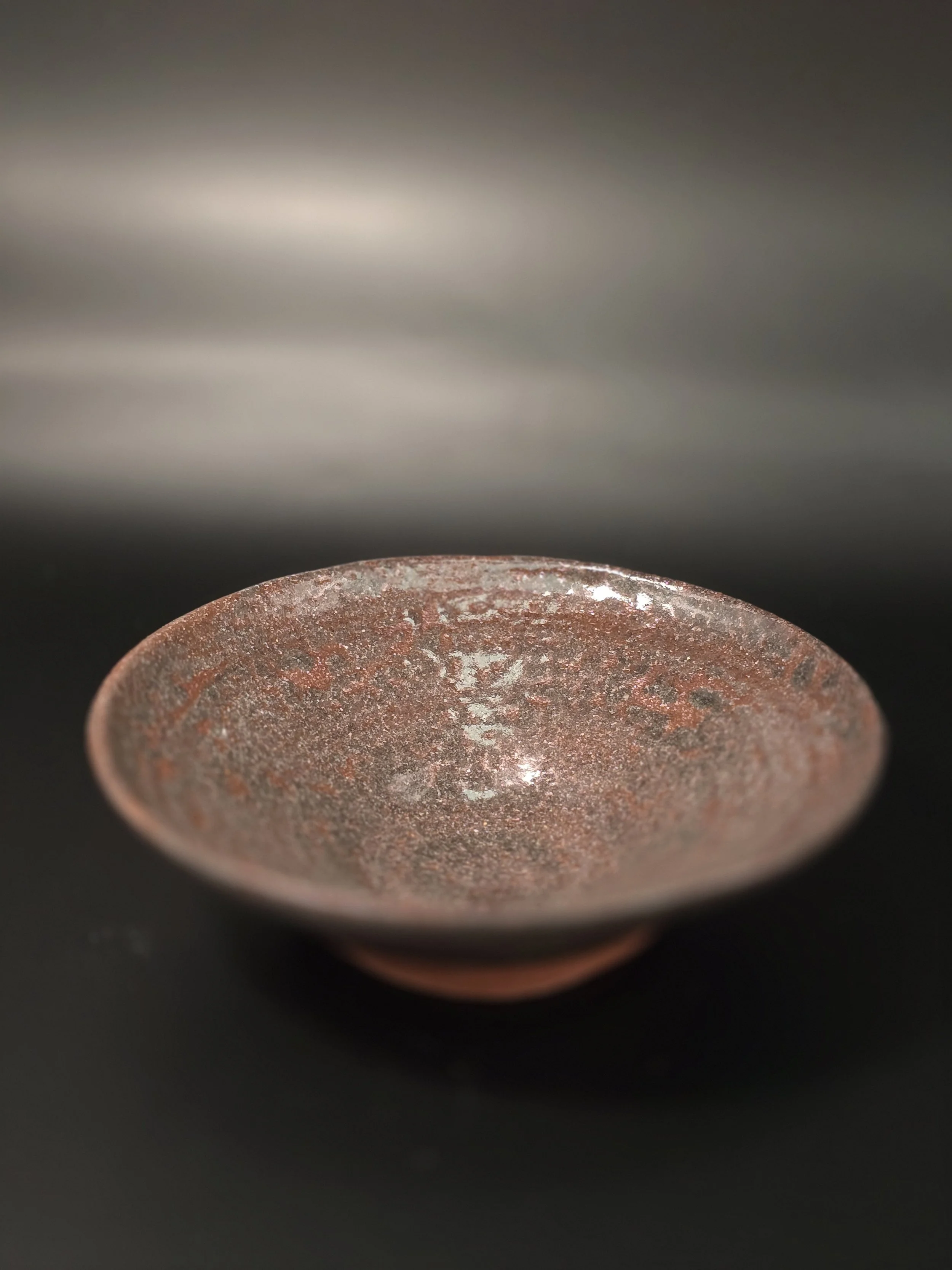 Close-up of a shallow, round, metallic bowl with a textured, reddish surface on a black background.