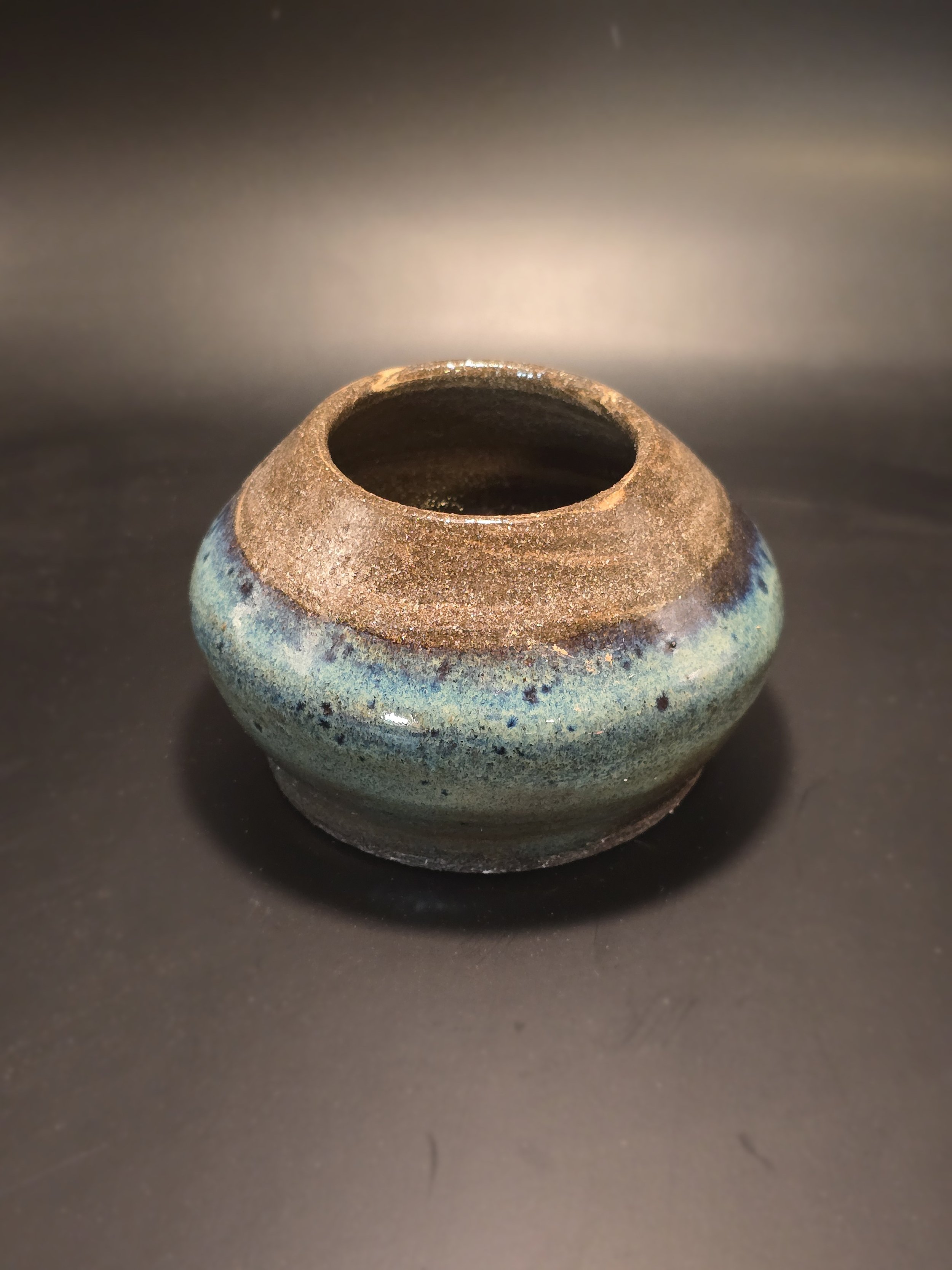 Small ceramic pot with a rounded shape, brown top, and blue-green glazed bottom, on a dark surface.