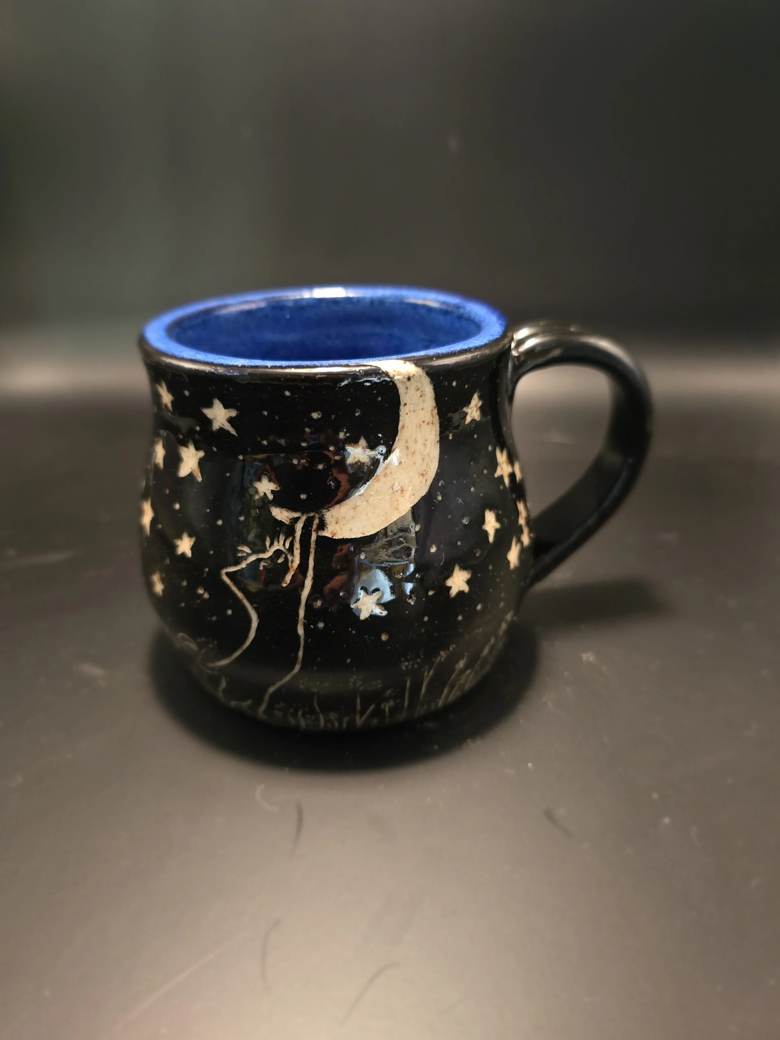 Black ceramic mug with space-themed design featuring a moon, stars, and a rocket ship; blue interior.