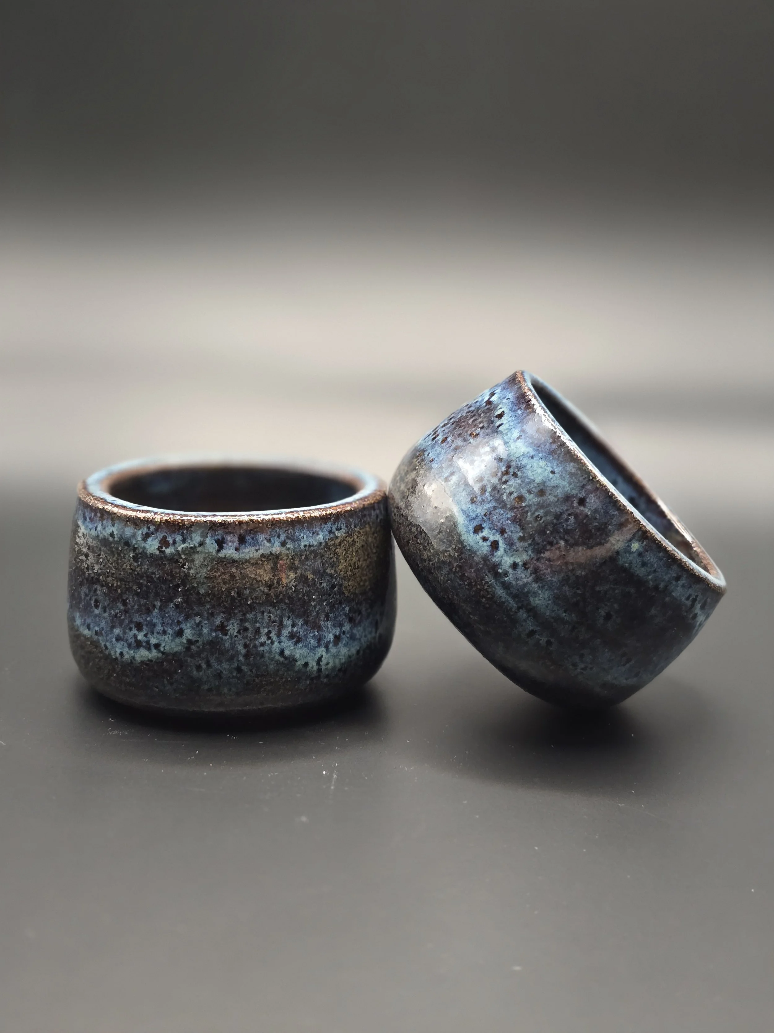 Two small ceramic cups with blue, black, and brown glaze, one cups appears to be leaning on the other, on a dark surface.
