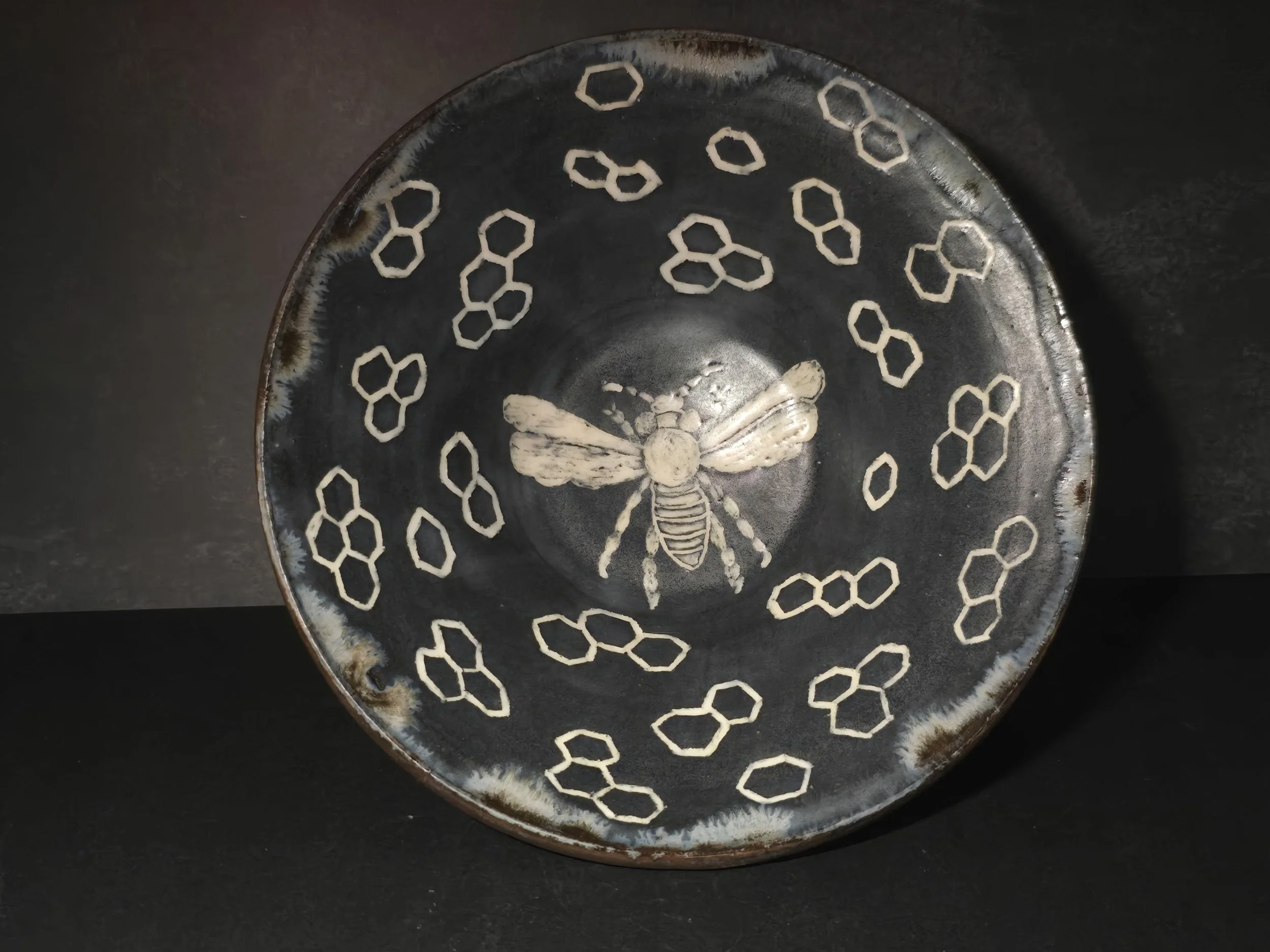 A dark ceramic plate with a bee illustration in the center, surrounded by hexagonal honeycomb pattern.
