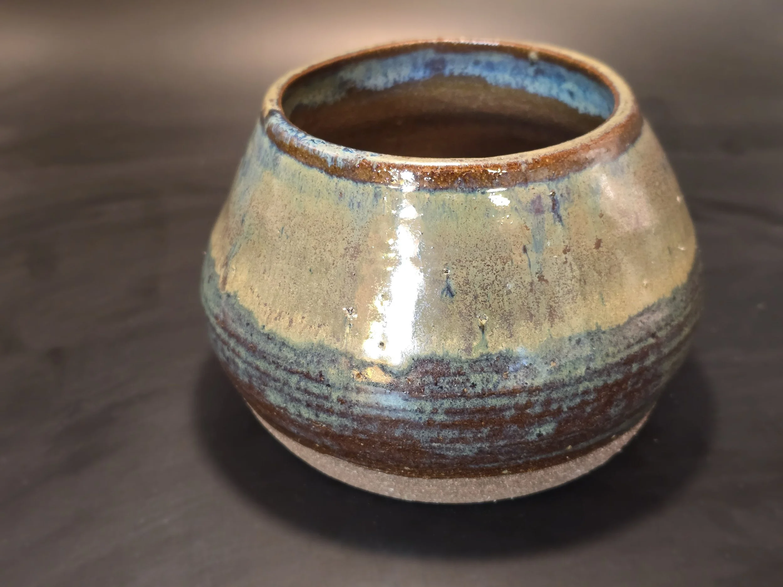 Ceramic pot with a glazed, multicolored surface, featuring earthy tones of brown, beige, and blue, placed on a dark wooden surface.