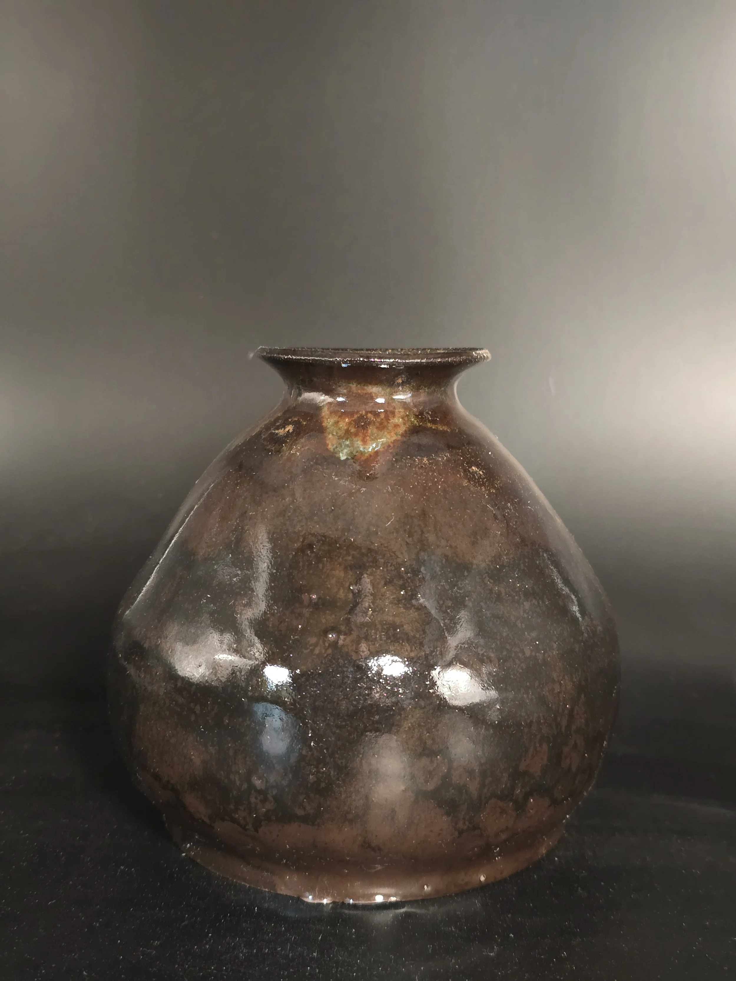 A small, rounded, dark brown ceramic vase with a narrow neck and slightly flared rim, sitting on a black surface against a gradient gray background.