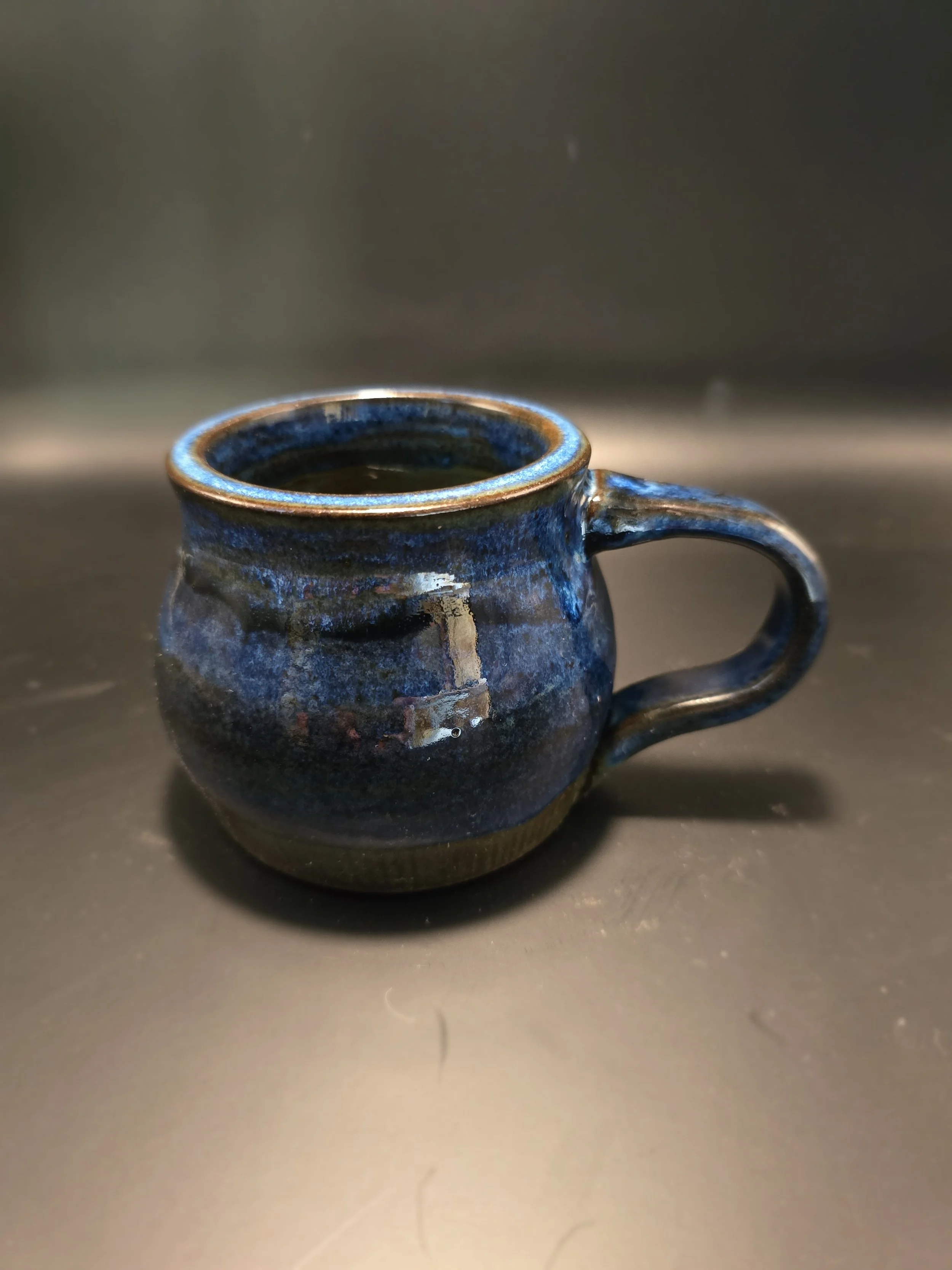 A small, dark blue ceramic mug filled with black coffee, placed on a metallic surface with a dark background.